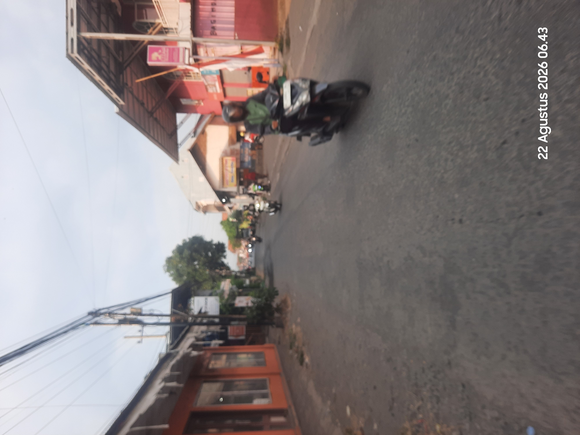 The image captures a street scene in Temenggungan, Indonesia, on August 22, 2026, at 06:43. The photograph, taken from a slightly elevated perspective, shows a paved road with a slight incline. On the left side of the road, there are several buildings with orange and white facades, featuring large glass windows. A few trees and streetlights are visible along the sidewalk. On the right side of the road, there are more buildings, including a prominent red structure with a modern, sloping roof and white accents. A small flag is attached to one of the buildings.
Several people on motorcycles are present on the road. In the foreground, a person wearing a helmet and a green jacket is riding a black scooter. Further down the road, other motorcycles are visible, some with riders and some without. The sky is overcast with a light gray hue, suggesting early morning or cloudy conditions. The overall atmosphere is that of a typical urban or semi-urban environment in Indonesia during the morning.