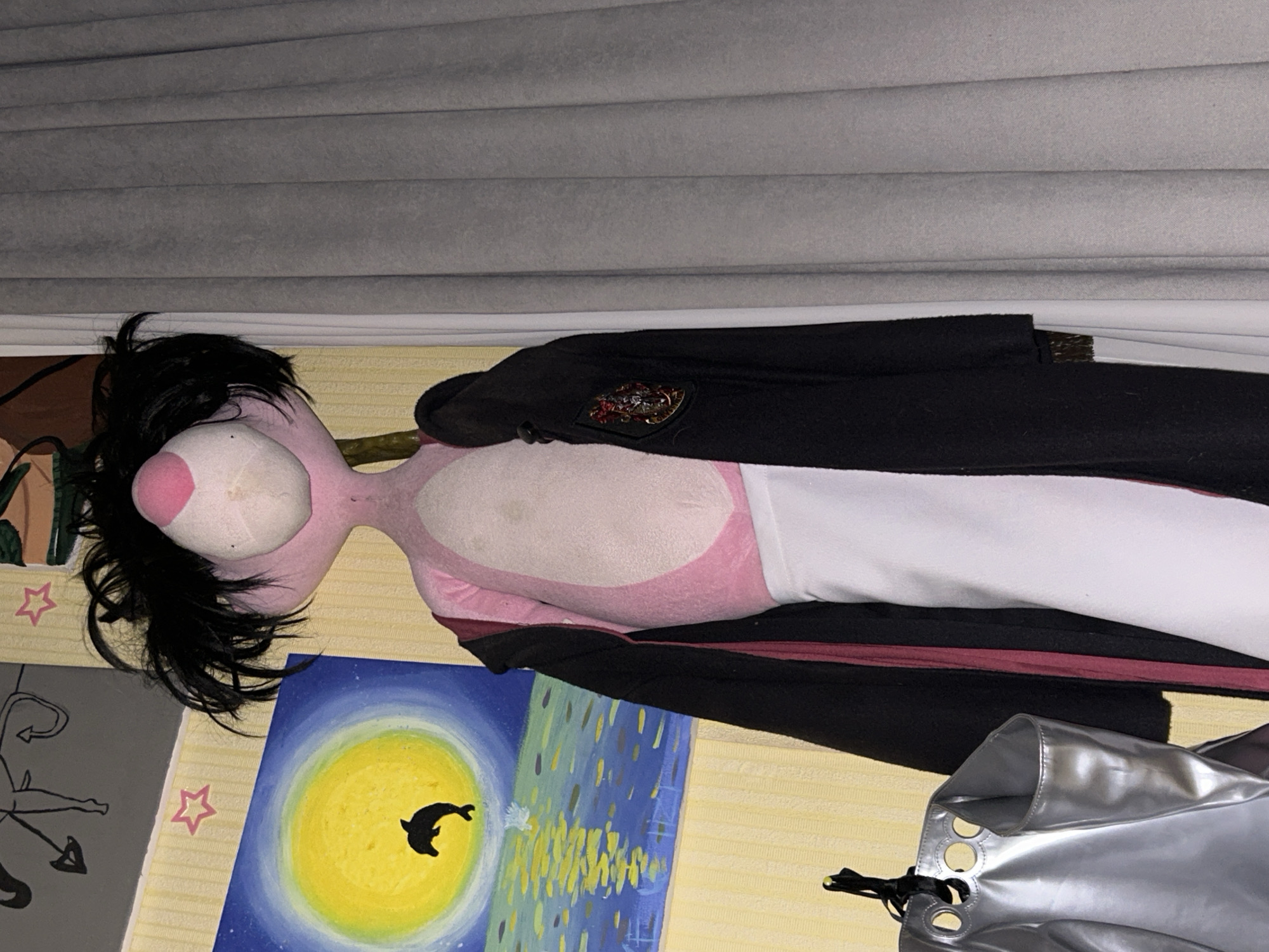 An indoor scene features a pink stuffed toy, likely a panther, dressed in a black robe with a Hogwarts emblem. The stuffed toy has black, messy hair and is positioned next to a silver tote bag hanging from a hook. Behind the stuffed toy, a painting depicts a yellow moon over a blue ocean with a jumping dolphin. The wall behind the painting is yellow and patterned. Gray curtains are visible to the right of the stuffed toy. The scene appears to be a bedroom or a decorated indoor space.
