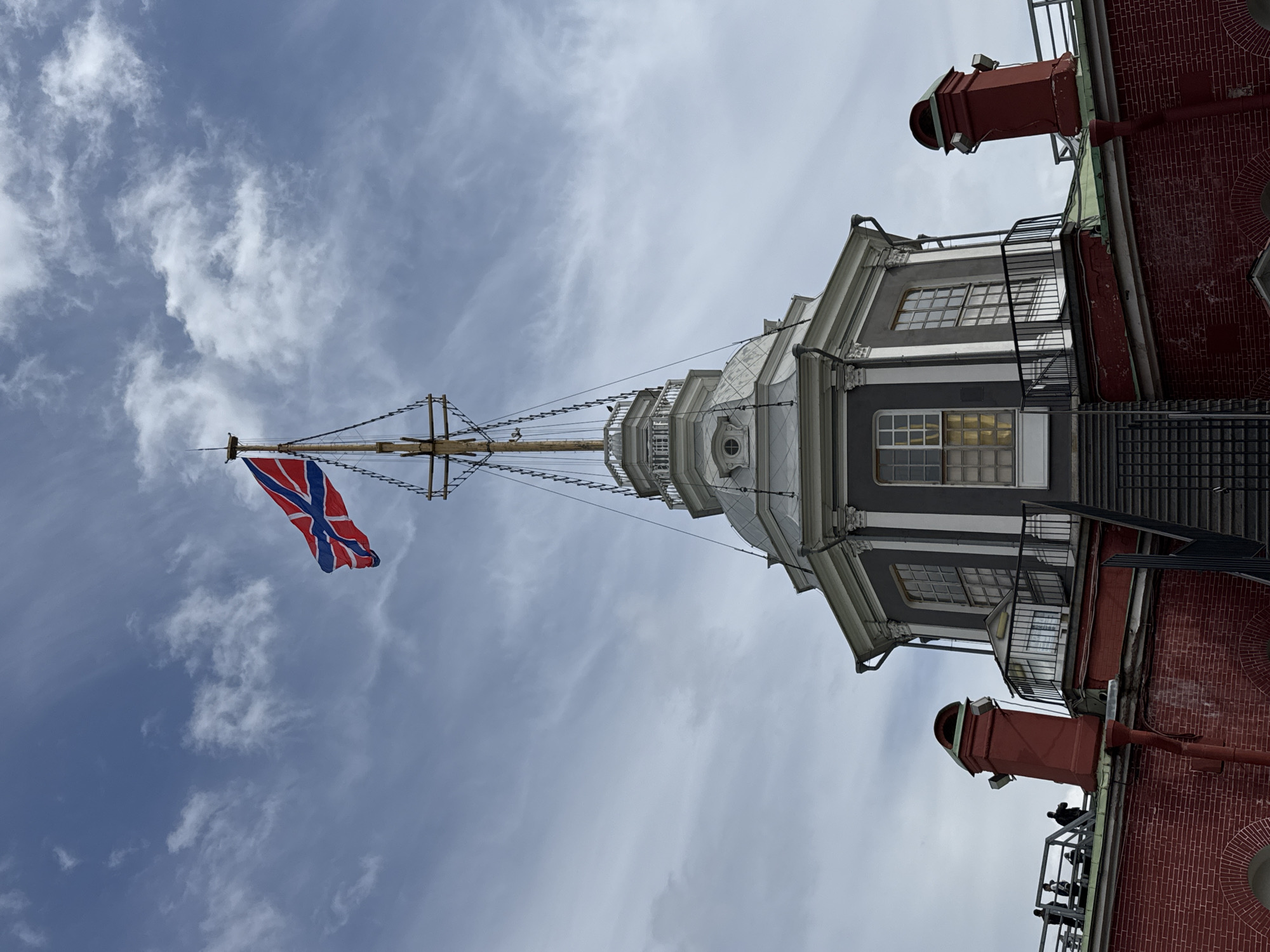 An outdoor, upward-looking photograph captures a distinctive architectural structure under a partly cloudy sky, with strong cues suggesting a historical naval location in Saint Petersburg, Russia.
The primary subject is a tall, slender wooden mast, intricately rigged with numerous ropes and lines, extending skyward. From its peak, a Russian St. Andrew's naval ensign unfurls gracefully in the wind. The flag is characterized by a blue saltire (diagonal cross) on a white field, with a subtle red outline bordering the blue arms.
The mast appears to rise from a multi-tiered, ornate building or superstructure. This central structure features several levels of grey and white façades, adorned with classical architectural details such as pilasters, cornices, and decorative grilles. Multiple windows, some rectangular and others circular (oculi or portholes), are visible, reflecting glimpses of light. Balconies with railings are also present on some levels.
To the right and below this central structure is a distinct red brick building. It showcases traditional brickwork and several large, multi-paned windows. Its roof is equipped with prominent, reddish chimney-like vents or pipes, some with distinct caps. A dark, metal external staircase or fire escape is mounted against the red brick wall, leading upwards. Faint dark shapes, likely pigeons, are perched on the roof of this red brick section.
The sky is a mix of light blue and soft white clouds, indicating daytime. There is no visible text in the image.