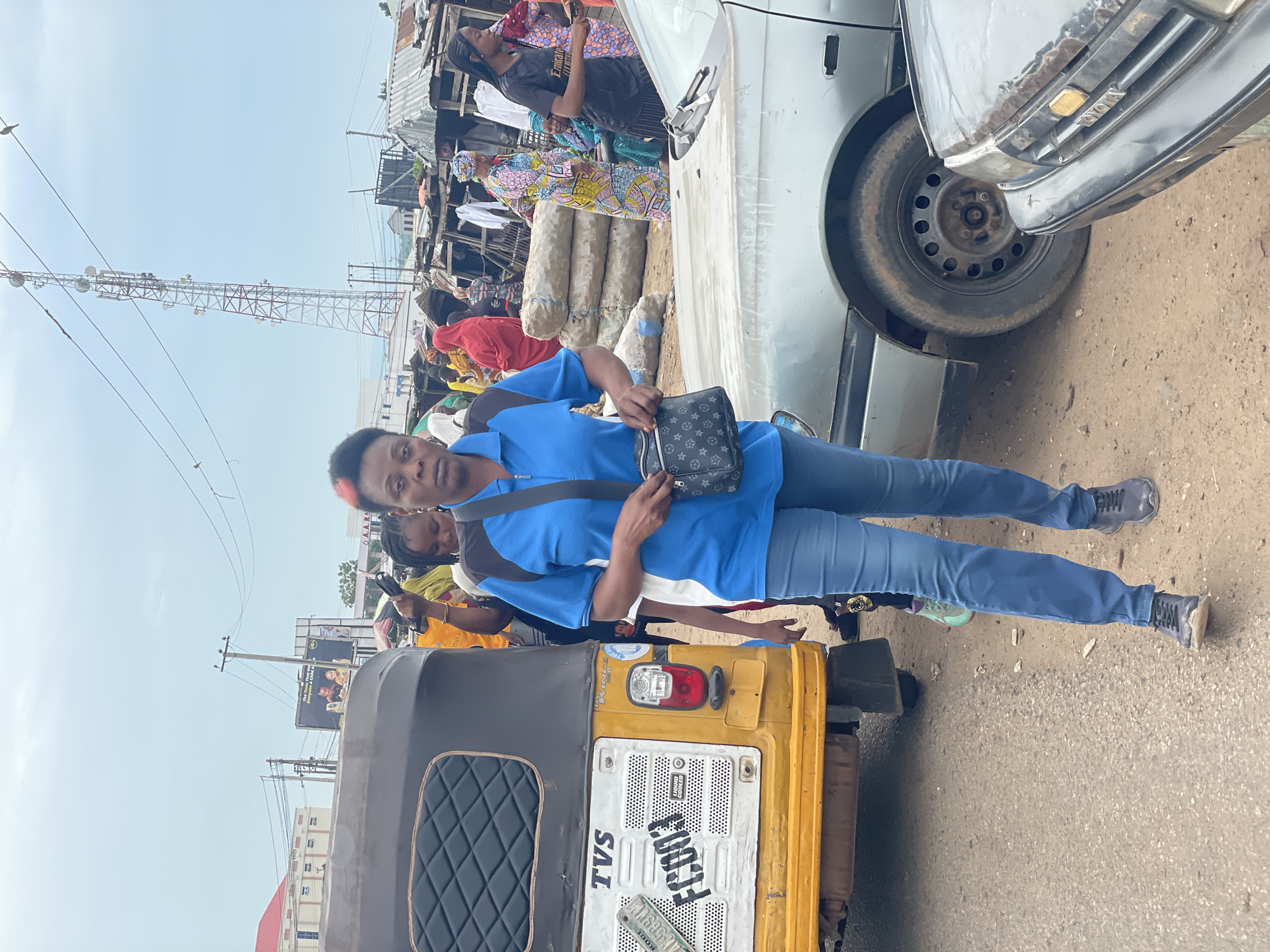 The image depicts a street scene in Lokoja, Nigeria. The focal point is a woman wearing a blue polo shirt and blue jeans, standing in the foreground and looking towards the camera. She is carrying a black purse with a Louis Vuitton pattern. Behind her, a yellow and grey autorickshaw, identified by the license plate "FC003", is visible. To the right of the autorickshaw, a silver sedan is parked. In the background, a bustling market environment is suggested by stalls, piles of goods, and numerous people, some of whom are wearing traditional clothing. A large telecommunications tower stands prominently in the sky above the market. The overall atmosphere is one of an active urban marketplace.