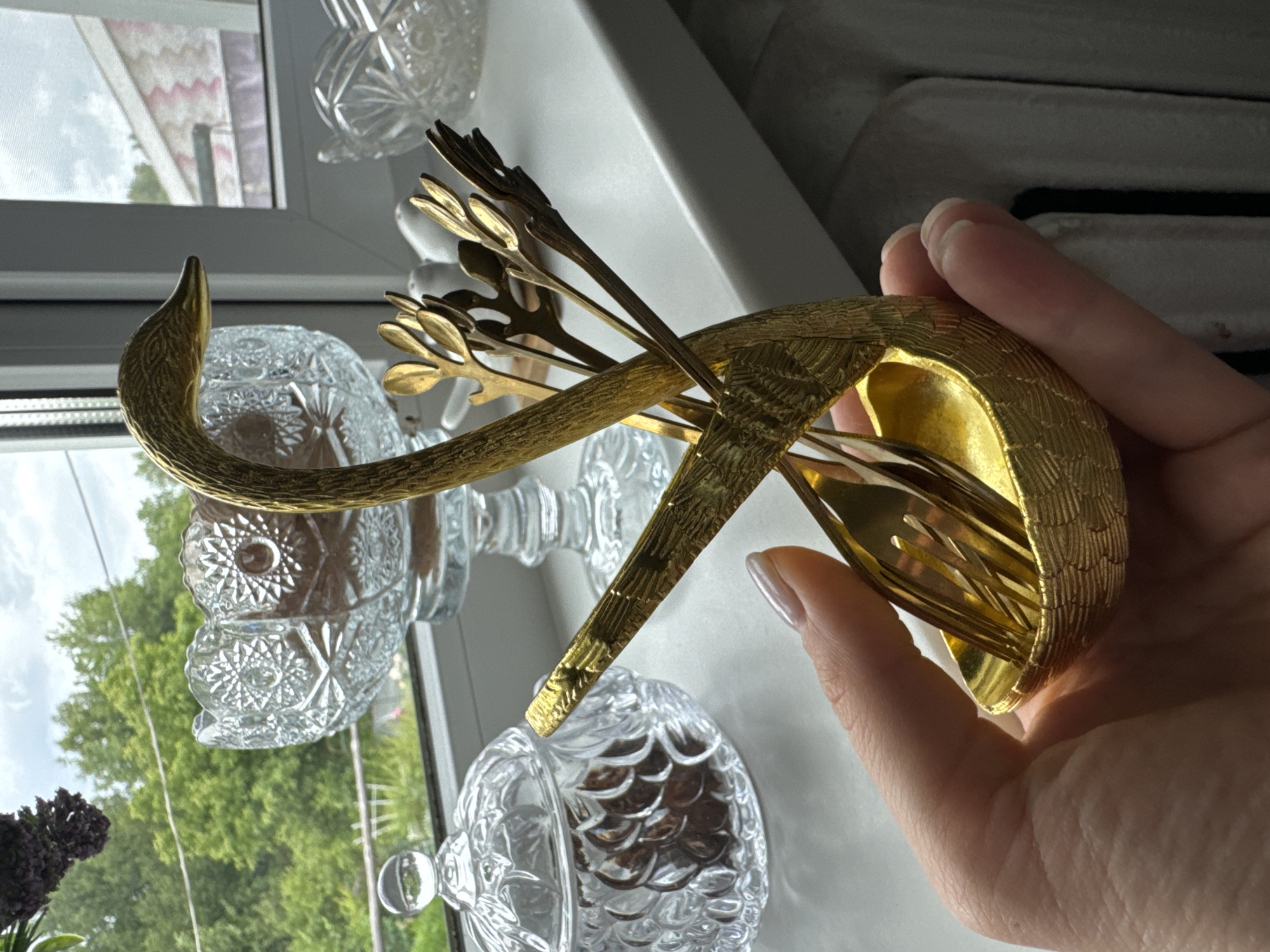 A hand with manicured nails holds a gold swan-shaped object, likely a decorative serving piece, filled with several gold forks. The swan's neck curves upwards, and its body is textured with a scale-like pattern. The background features a windowsill with several clear crystal decorative objects, including a faceted bowl and a smaller, intricately patterned bowl, placed in front of a window. Through the window, a slightly overcast sky and lush green trees are visible, suggesting it is daytime. The overall scene appears to be indoors, in a home setting, with a focus on the decorative gold swan and crystalware. No text is visible.