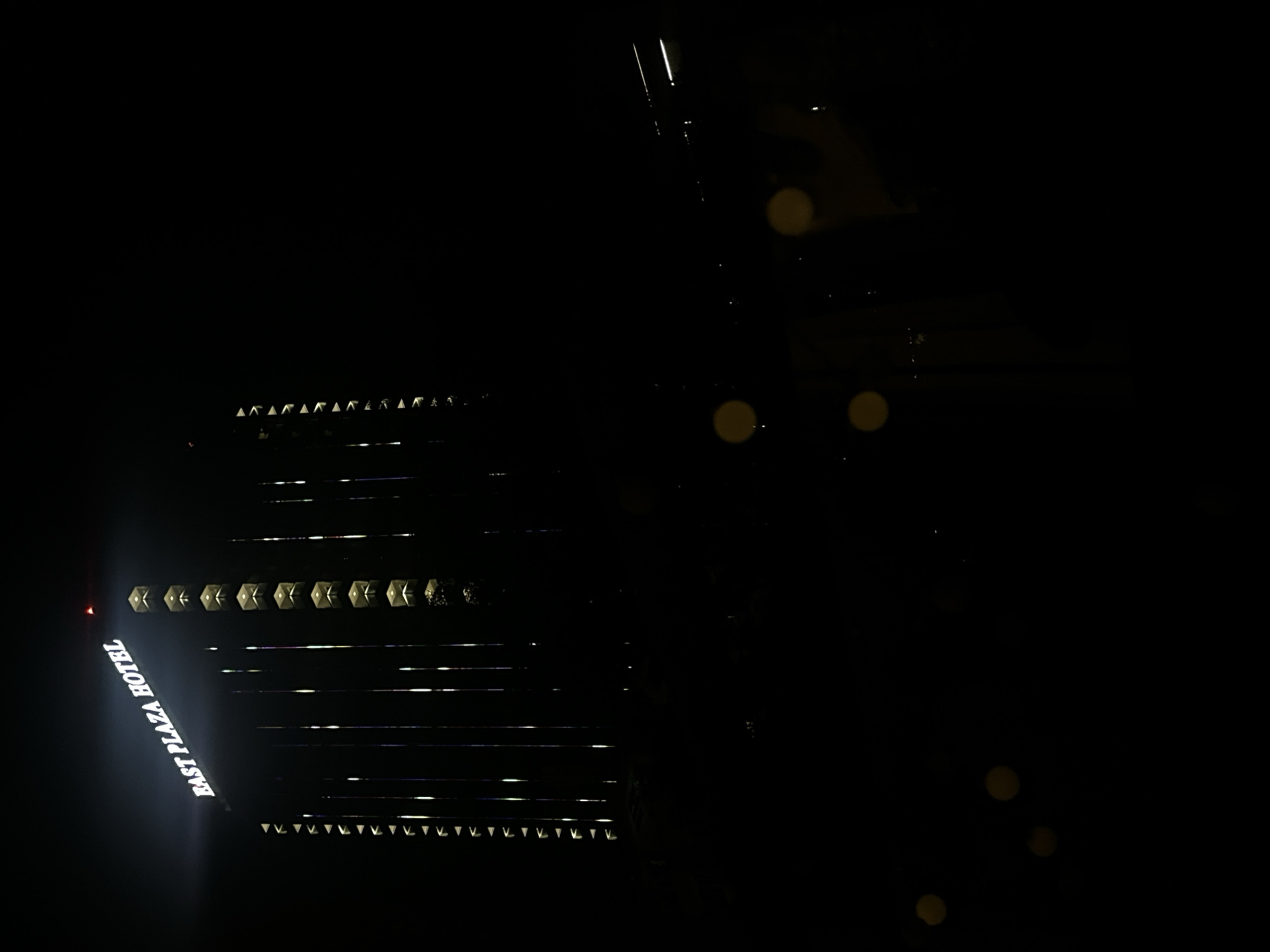 A nighttime exterior shot shows the lit-up "EAST PLAZA HOTEL" sign on a tall building. The hotel's facade is dark, with vertical light accents and rows of illuminated windows. The surrounding environment is black, suggesting it's a dark night or the image is primarily focused on the building. There are some out-of-focus light orbs in the lower portion of the frame, possibly from streetlights or vehicles. There are no people or specific actions visible. The scene is likely in Sofia, Bulgaria, given the mention of the city.