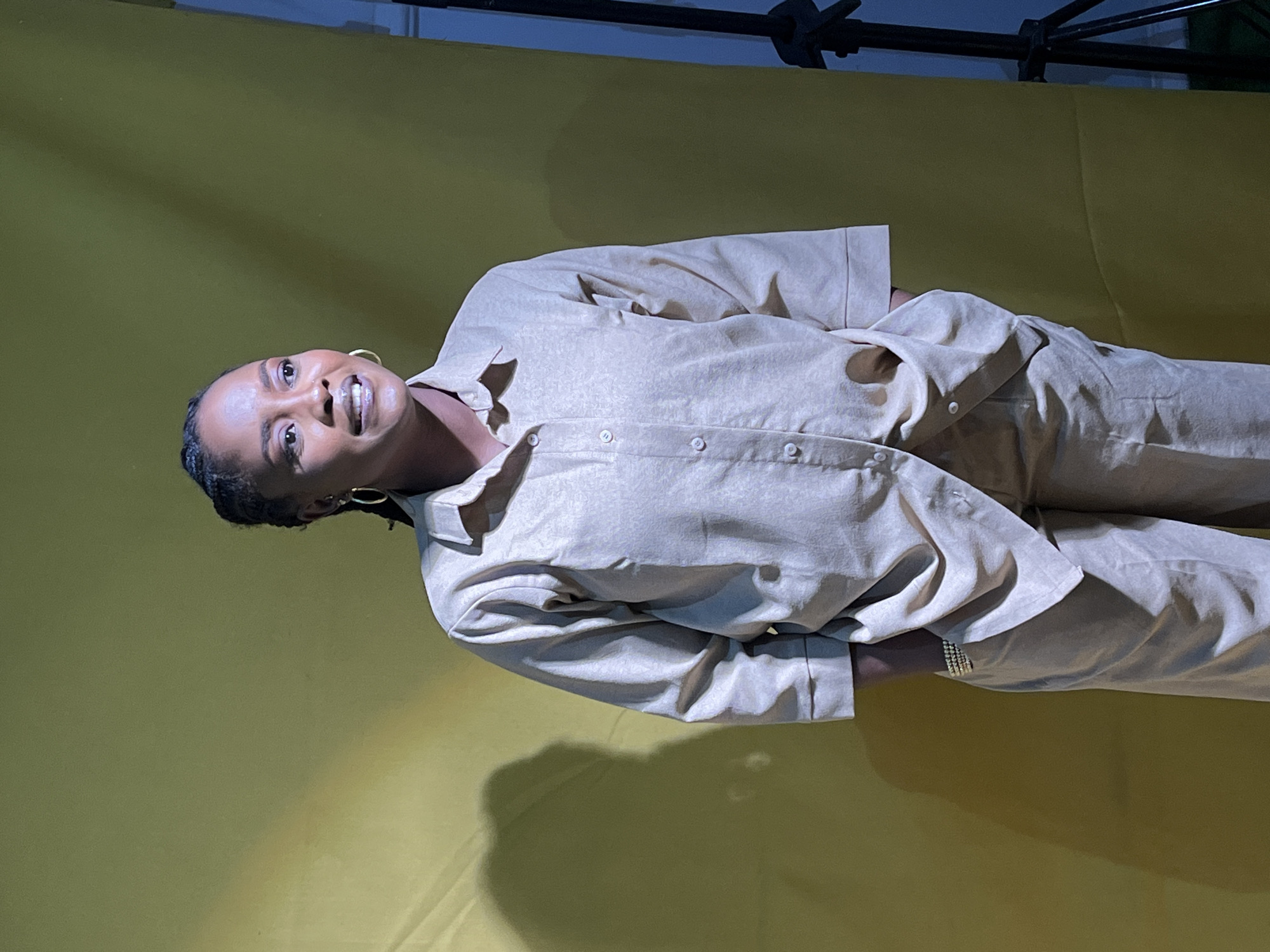 A woman with braided hair and gold hoop earrings stands against a yellow backdrop. She is wearing a light beige collared shirt with buttons down the front and matching pants. Her left hand is tucked into her pocket, revealing a bracelet. The lighting creates a shadow on the left side of the backdrop, and a tripod is visible on the right. The scene appears to be a studio or indoor setting. There is no specific information or visual cues in the image to suggest its location in Barakin Akawo, Nigeria.
