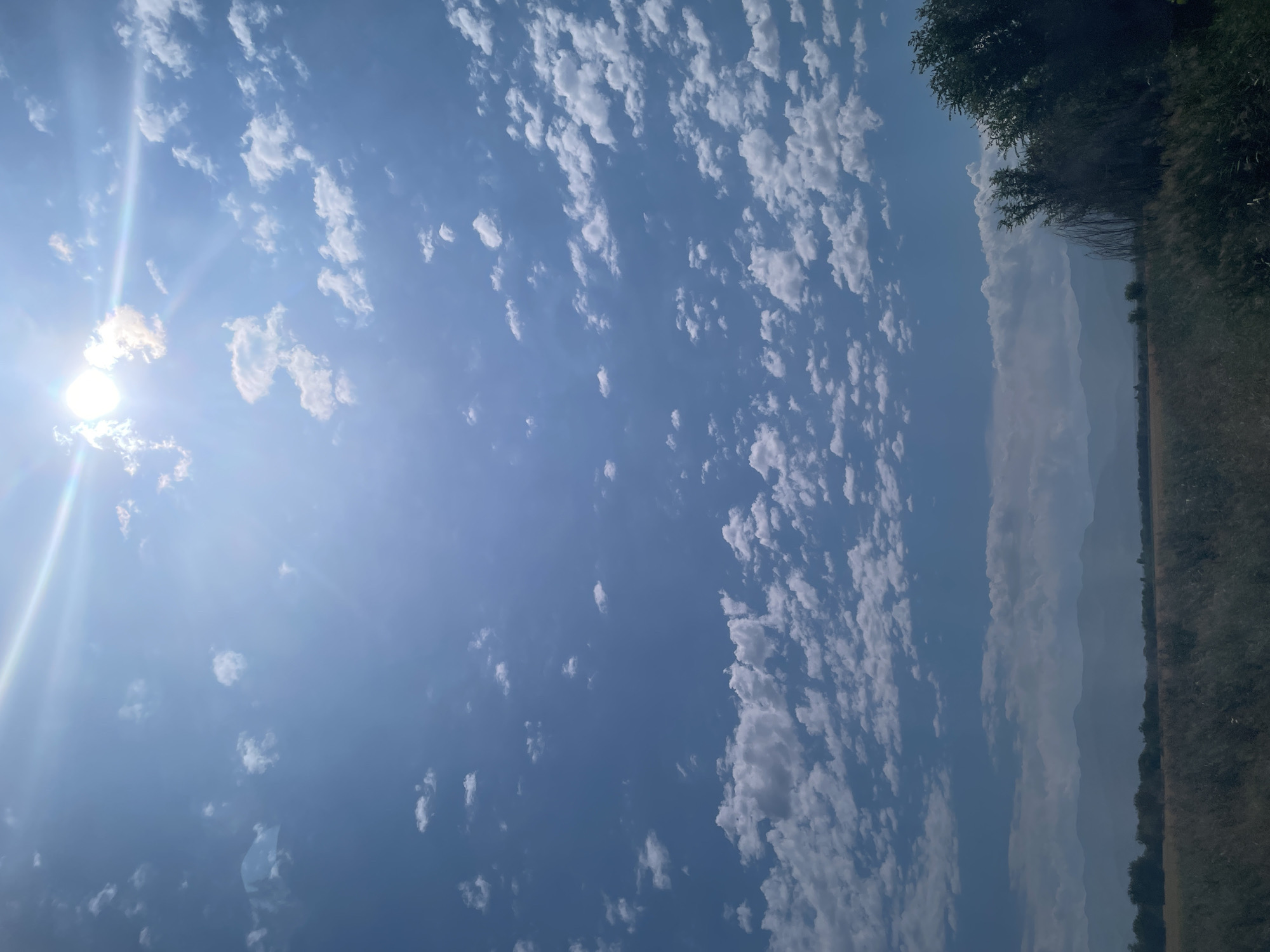 A wide shot captures a vast blue sky dotted with scattered white clouds on a sunny day. The sun, positioned in the upper right quadrant, casts a bright glare. Below the sky, a landscape unfolds with hazy mountains in the distance, a line of trees and vegetation, and a field of dry grass and plants in the foreground. The scene appears to be an outdoor, natural environment, possibly in a rural area or on the outskirts of a city. The overall atmosphere is serene and depicts a clear day with fair weather. The location is stated to be in the city of Ақбастау, Kazakhstan.