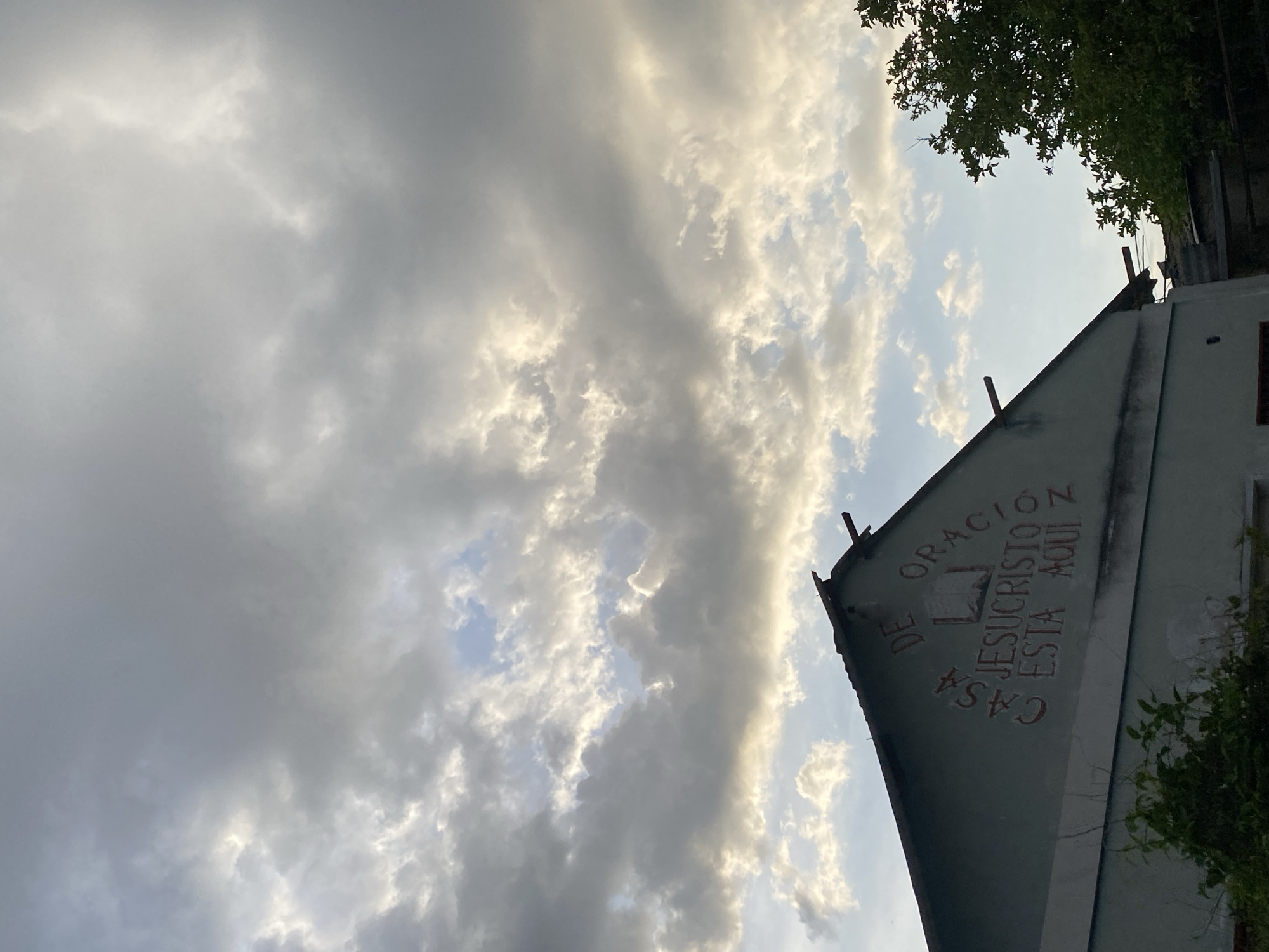 The image shows a building in Tinaquillo, Venezuela with the words "Casa de Oracion Jesucristo Esta Aqui" painted on its side. The building appears to be a simple structure with a peaked roof. The sky is cloudy, with some hints of sunlight breaking through. There are trees visible in the upper right corner of the image. The image is taken from a low angle, looking up at the building and sky.