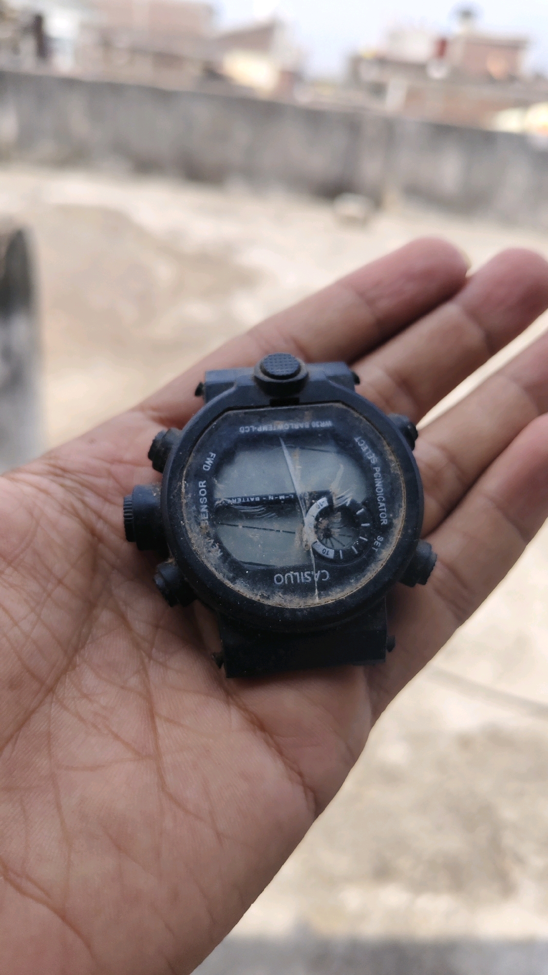 In Thikri, India, a black, digital wristwatch with a cracked screen rests on a person's open palm, which is held in front of a blurred background of buildings and a dusty, concrete surface. The watch displays some visible text, including "CASILUO," "SENSOR," "FWD," and other unreadable markings. The watch is also dirty, suggesting it may have been exposed to the elements. The person's hand shows visible lines and texture, indicating an adult hand. The overall scene is an outdoor setting, possibly a rooftop or open area, given the background.