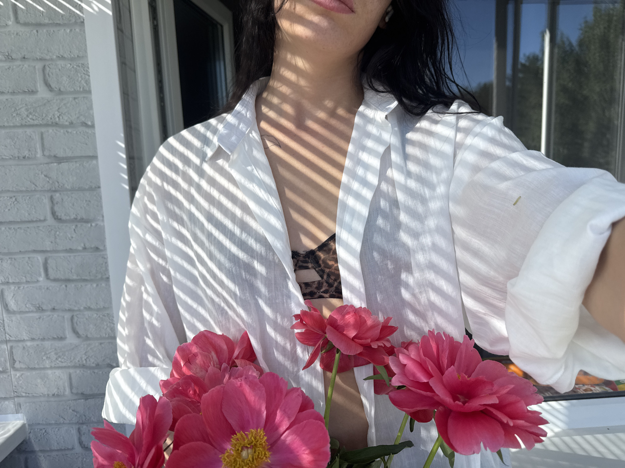A medium shot captures a person from the chest up, outdoors, with a white brick wall on the left and a window with trees visible through it on the right. The person, wearing a white unbuttoned shirt over a leopard-print bra, is holding a bouquet of pink peonies. Sunlight casts striped shadows across the person's chest and the white shirt. The person's dark hair is visible around their shoulders.