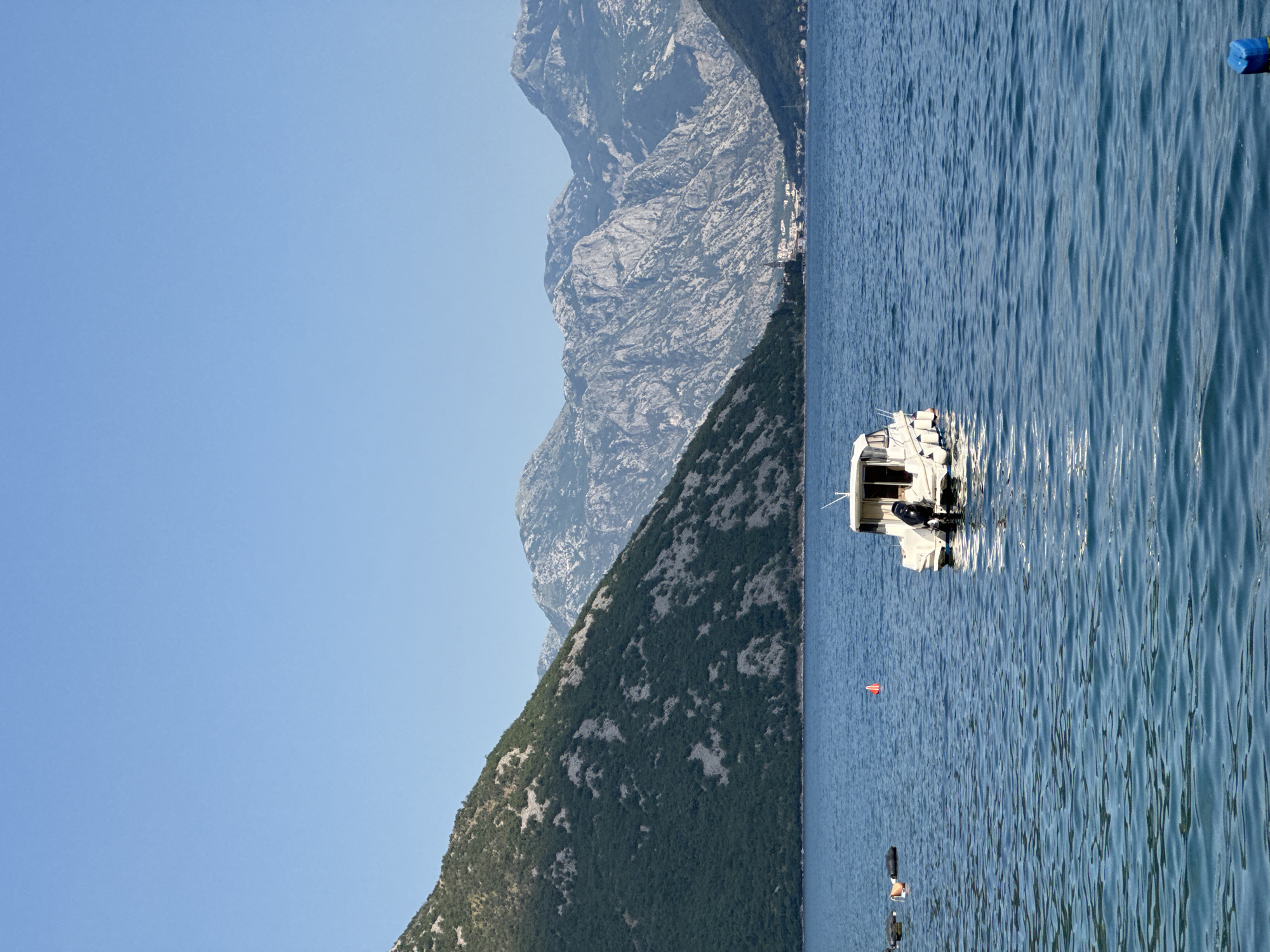 In a serene, sun-drenched scene, a calm, blue body of water stretches towards a backdrop of majestic, rugged mountains. The mountains, covered in lush green vegetation on their lower slopes and rocky grey formations higher up, rise dramatically against a clear, pale blue sky. In the foreground, a small white motorboat with a canvas canopy and an outboard motor is anchored in the water. To the left of the boat, several black buoys are visible, along with a small orange conical buoy. The overall atmosphere is one of peaceful tranquility, evoking a sense of being in a natural, secluded haven. The location is identified as Strp, Montenegro.