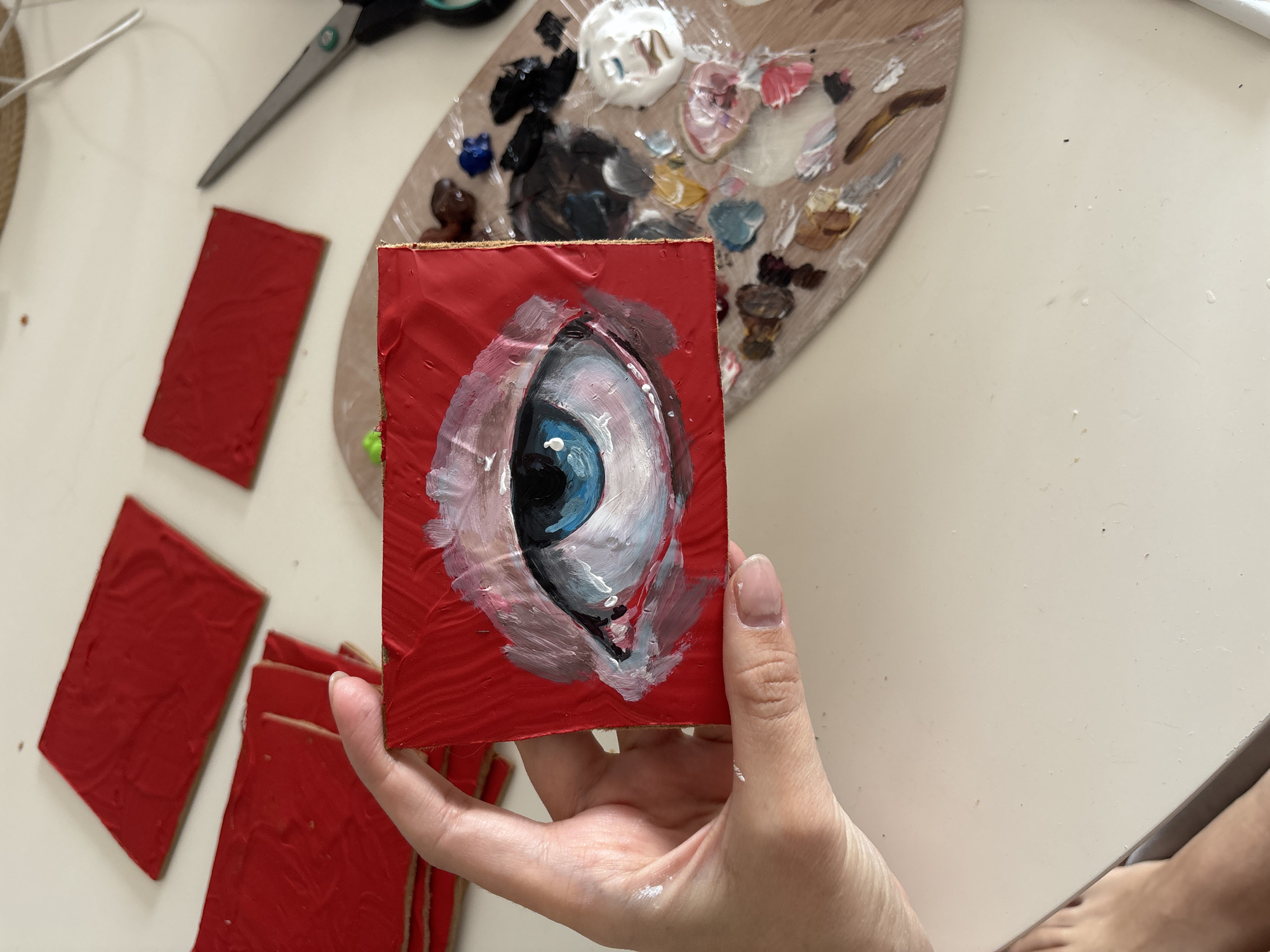 The image depicts a person holding a small, square painting of an eye with a blue iris and white highlights on a red background. The painting appears to be done in an impressionistic style with visible brushstrokes. Several other red painted squares are lying on a white surface to the left of the painting. A painting palette with various blobs of color is located above the artwork, along with a pair of black-handled scissors in the upper left corner. The person's hand is visible holding the painting, and a glimpse of their foot is seen in the lower right corner. The environment appears to be a crafting or art studio. There is no indication this picture has a location in the city of Krasnoyarsk, Russia.