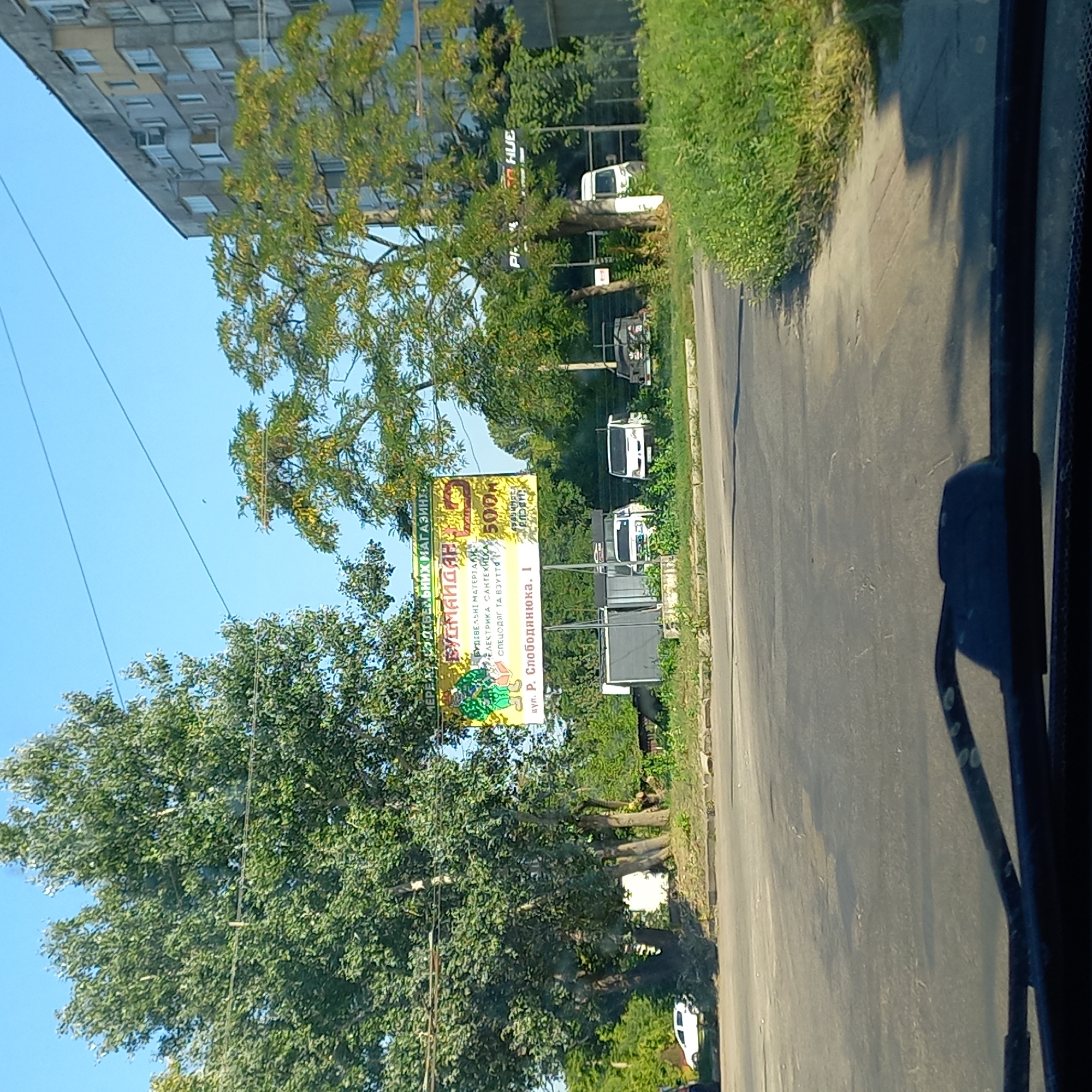 The image captures a street scene in Kropyvnytskyi, Ukraine, on a clear, sunny day. In the foreground, the dashboard and windshield wipers of a vehicle are visible. Beyond the vehicle, a paved road curves to the left. On the right side of the road, there is a dense area of green grass and foliage. Further back, a large billboard with yellow and green coloring is prominently displayed. The billboard features text in Ukrainian, with a large graphic of two cartoonish figures wearing overalls. Below this graphic, more text indicates "БУДІВЕЛЬНІ МАТЕРІАЛИ" (building materials), "ЕЛЕКТРИКА САНТЕХНІКА" (electricity, plumbing), and "СПЕЦОДЯГ ТА ВЗУТТЯ" (work clothes and footwear). The street address "вул. Р. Слободянюка, 1" (R. Slobodianiuk St., 1) is also visible.
Behind the billboard, a series of white vans are parked. To the right of the vans, a multi-story, gray residential building with multiple windows can be seen. A portion of a sign with "PH" and "HUE" is visible on the right side of the image, near a tree and the residential building. Two tall trees with full green foliage stand on the left side of the frame, partially obscuring the view of the residential building and the sky. Another tree with lighter green foliage is positioned to the right of the billboard. A white passenger car is parked on the far left side of the image, on a grassy embankment. The overall environment suggests a commercial or semi-industrial area within the city.