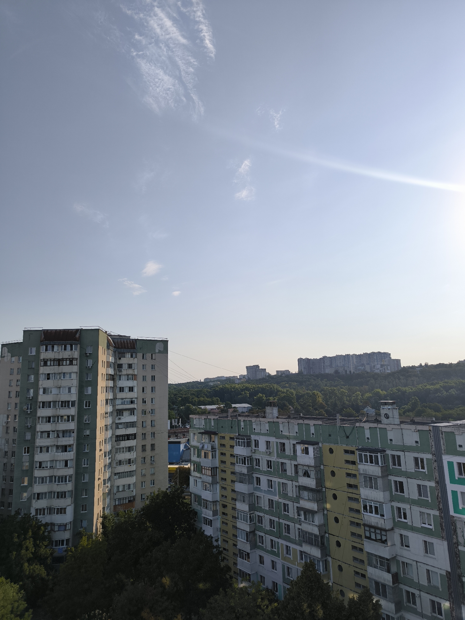 The image shows a panoramic view of an urban landscape with a clear sky and a few wispy clouds. In the foreground, several apartment buildings are visible, characterized by their repetitive architecture, balconies, and window arrangements, common in Soviet-era housing blocks. The buildings are painted in muted colors, with accents of green, yellow, and grey. Trees and greenery are interspersed between and around the buildings, suggesting a residential area with parks or green spaces. In the background, more distant buildings and a densely forested hill can be seen. The sun is positioned in the upper right corner, casting a lens flare across the sky. The scene is a typical depiction of a residential district in Chișinău, Moldova, likely captured from a higher vantage point, such as another building or elevated terrain.