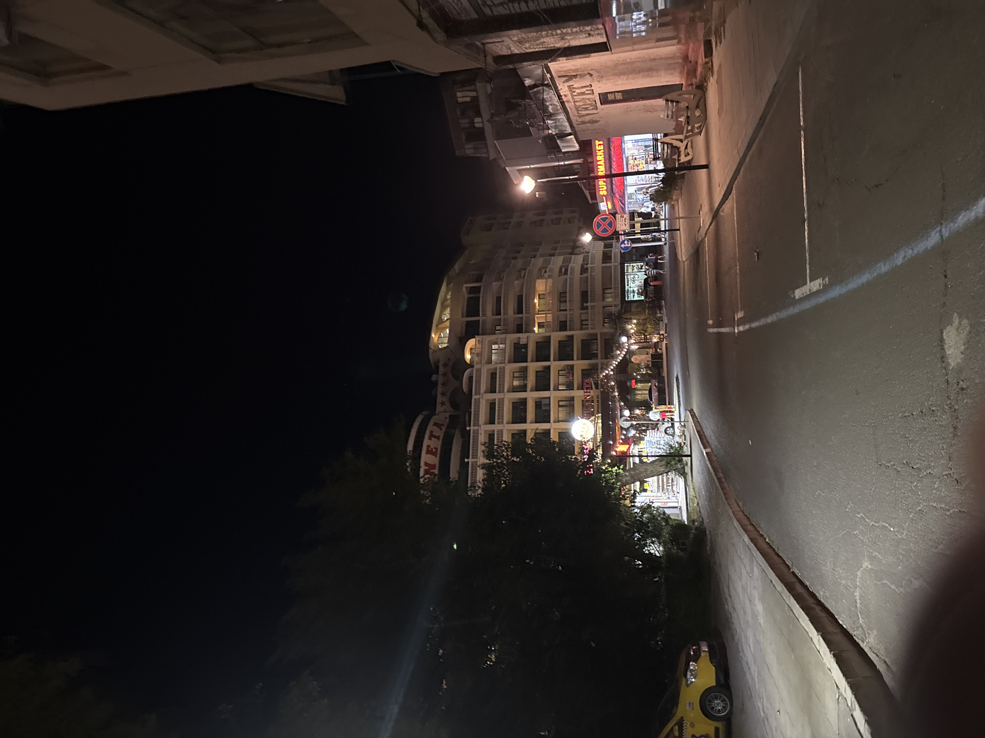 The image depicts a street scene at night in Nesebar, Bulgaria, likely near a resort or beach area. On the left, a yellow taxi is parked along a sidewalk. Dominating the background is a large, multi-story hotel complex with illuminated balconies and a sign that reads "PLANETA". To the right of the hotel, a row of storefronts is visible, including a "SUPERMARKET" with bright red signage and a store labeled "VELVET". Streetlights illuminate the paved road and sidewalk, where a few people can be seen walking. The overall atmosphere is that of a commercial street with tourist-oriented businesses operating at night.