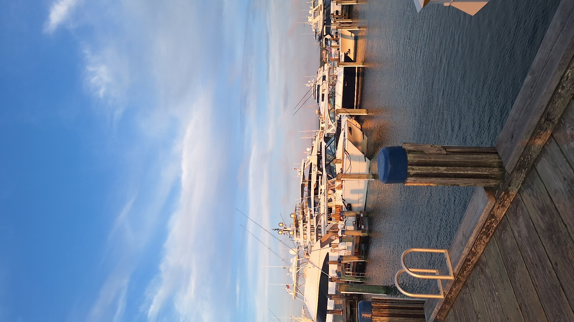 In Nantucket, Massachusetts, USA, a collection of luxury yachts and smaller boats are moored at a dock. The wooden dock is in the foreground, with weathered planks and a metal ladder extending into the water. Several pilings, some with blue caps, mark the edge of the dock. The water is calm, reflecting the sky and the boats. The boats are docked in a line, with the largest yacht in the background, featuring multiple decks and prominent fishing rods. Smaller, sleeker vessels are in the mid-ground. The sky above is a bright blue with scattered white clouds, illuminated by what appears to be the warm light of either sunrise or sunset. There are no people visible in the image.