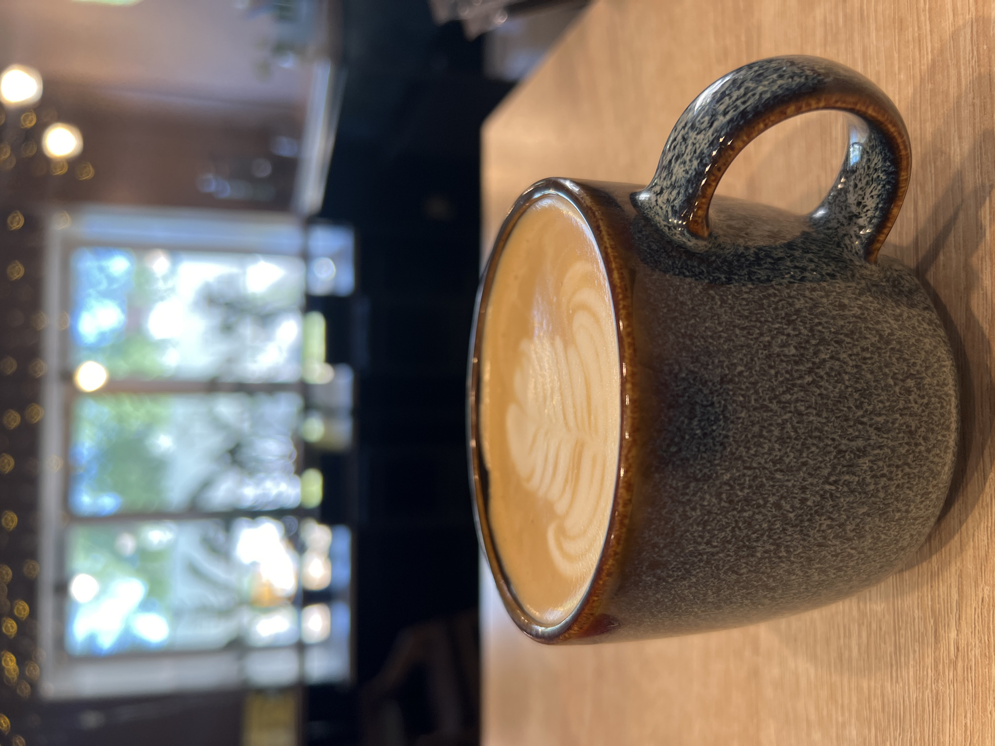 A close-up, slightly canted, indoor shot focuses on a coffee mug filled with latte art. The mug is a mottled blue-grey with a brown rim and handle, resting on a light-colored wooden surface. The latte art is a light brown color with a leaf pattern.
In the background, out of focus, is a window with a view of trees and possibly other buildings. To the left of the window, blurred lights are visible, giving a bokeh effect.
The location in the city of Chaikovsky, Russia, cannot be determined from the provided image. There are no identifiable markers to confirm the location.