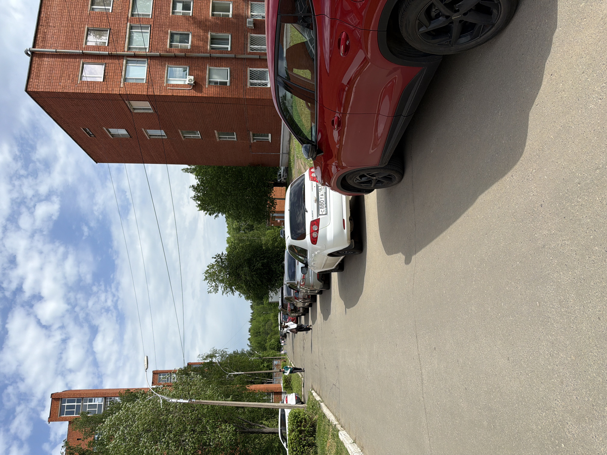The image shows a street in Izhevsk, Russia, lined with parked cars. A tall brick apartment building is on the left side of the street, with trees and another lower brick building visible in the background. Power lines run along the street. Several people are walking along the street. A red car and a white Chevrolet Lacetti are prominently parked in the foreground. The license plate of the Lacetti is visible as "C 880 TA 18". The street is paved, and the weather appears sunny.