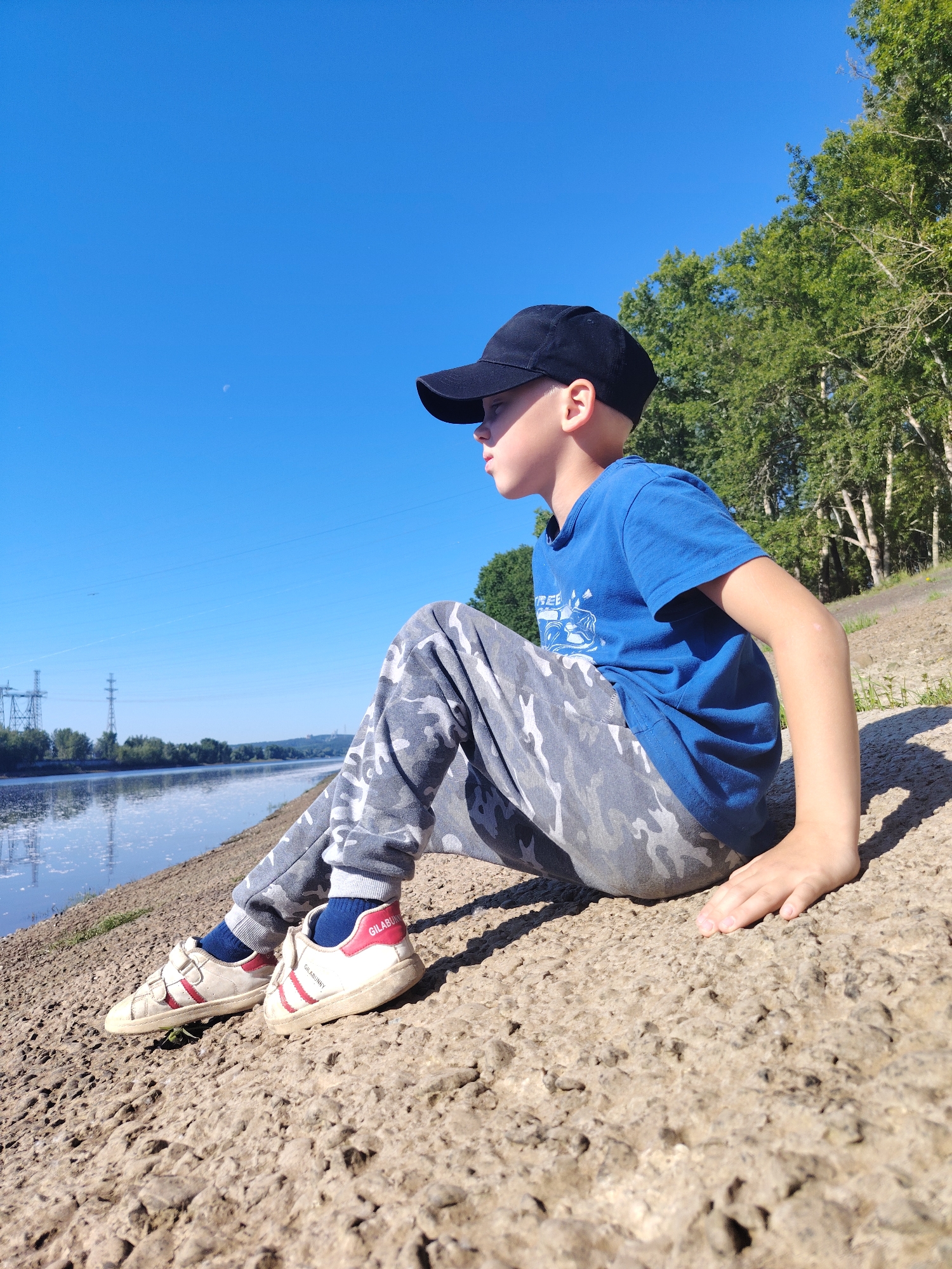 In Chaikovsky, Russia, a young boy in a blue t-shirt and camouflage pants sits on a rocky embankment beside a calm river under a clear blue sky. He wears a black baseball cap and white sneakers with red stripes. The river reflects the sky and a distant treeline with power lines. The scene is bathed in sunlight, casting shadows from the boy and the surrounding trees.
