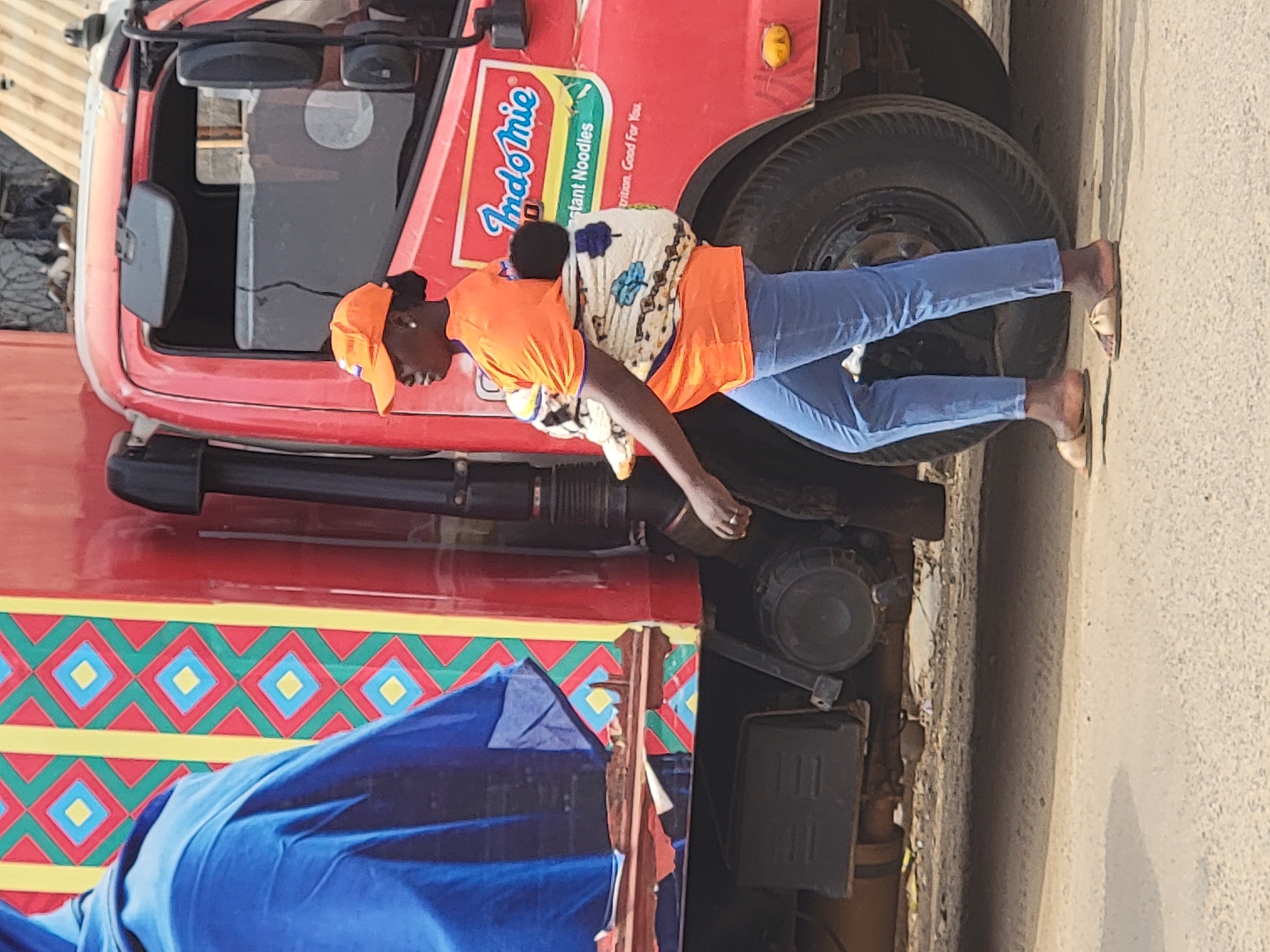 A woman wearing an orange shirt, blue jeans, and a cap is carrying a child on her back. She is standing next to a red Indomie truck. The truck is decorated with colorful geometric patterns and a blue tarp. The location is Maiburiji, Nigeria.
