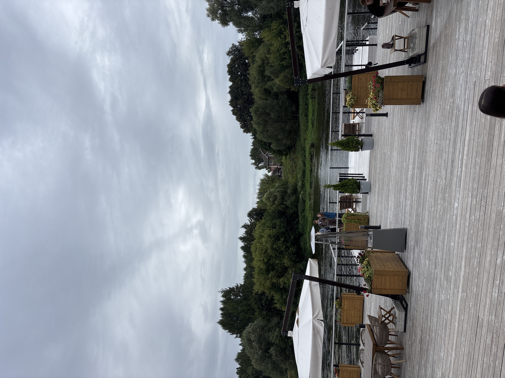 The image depicts an outdoor seating area on a wooden deck overlooking a body of water, likely a river, on an overcast day. The deck is furnished with several tables and chairs, two large white umbrellas, planters with flowers, and a patio heater. In the background, across the water, is a dense line of green trees and vegetation, with a few houses visible further back. Three people are standing on a small pier extending into the water. Another person is seated at a table on the deck in the lower right corner, and the back of a person's head is visible in the foreground. The overall atmosphere is calm and serene. This scene is likely located at a restaurant or cafe along the Moskva River in Zvenigorod, Russia, a city known for its natural beauty and historical sites.