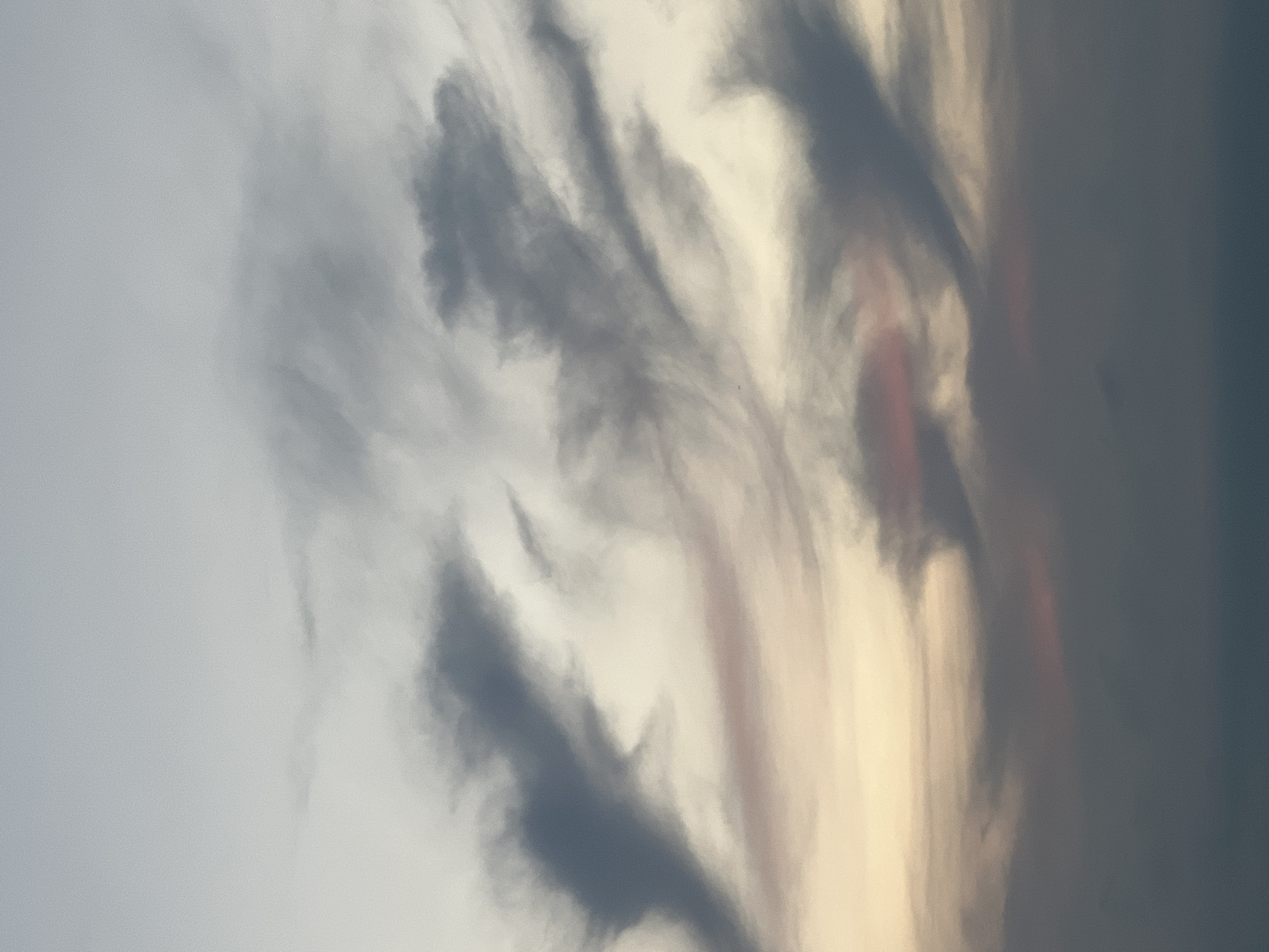 An image of wispy, gray clouds against a light sky. The clouds are textured and appear to be moving, with some streaks of pink and orange visible at the bottom of the frame where the sky transitions to a darker hue. The overall impression is that of a sunset or sunrise.