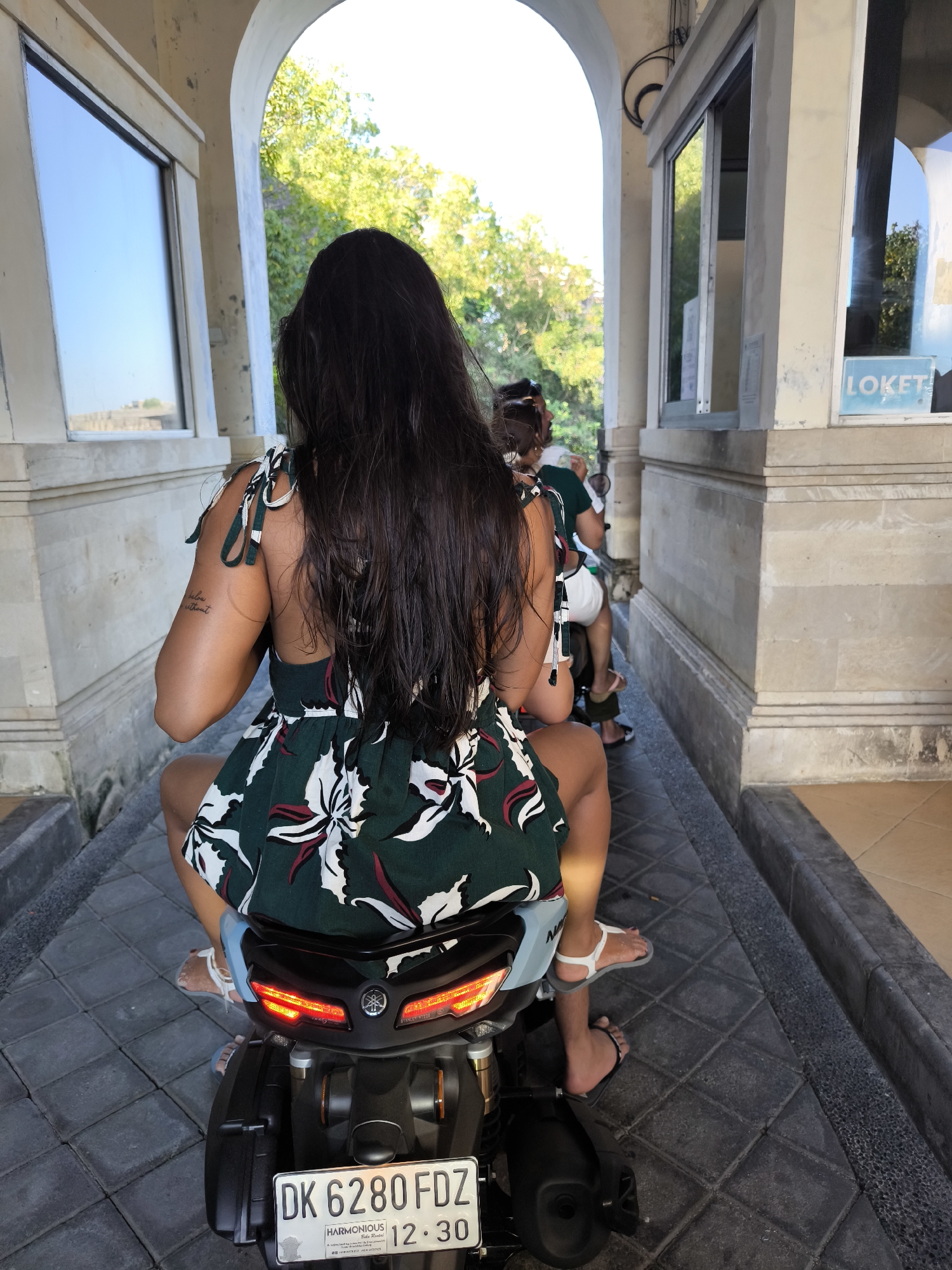 In Kuta Selatan, Indonesia, two individuals are seated on a scooter, preparing to pass through an arched entryway that appears to be a toll booth or entrance to a facility. The primary subject, a woman with long, dark, wet hair, is in the foreground, facing away from the camera and riding on the back of the scooter. She is wearing a green and white floral print dress with tie straps on the shoulders. Her legs are visible, and she is wearing white sandals. The scooter's license plate reads "DK 6280 FDZ" and displays text that appears to be related to "HARMONIOUS Ride Rental." Behind her, another person, partially obscured, is driving the scooter, wearing a green shirt and shorts. The environment features stone paving and a light-colored arched structure overhead, with greenery visible through the opening. The scene suggests movement and transition, likely entering or exiting an area that requires payment or a pass.
