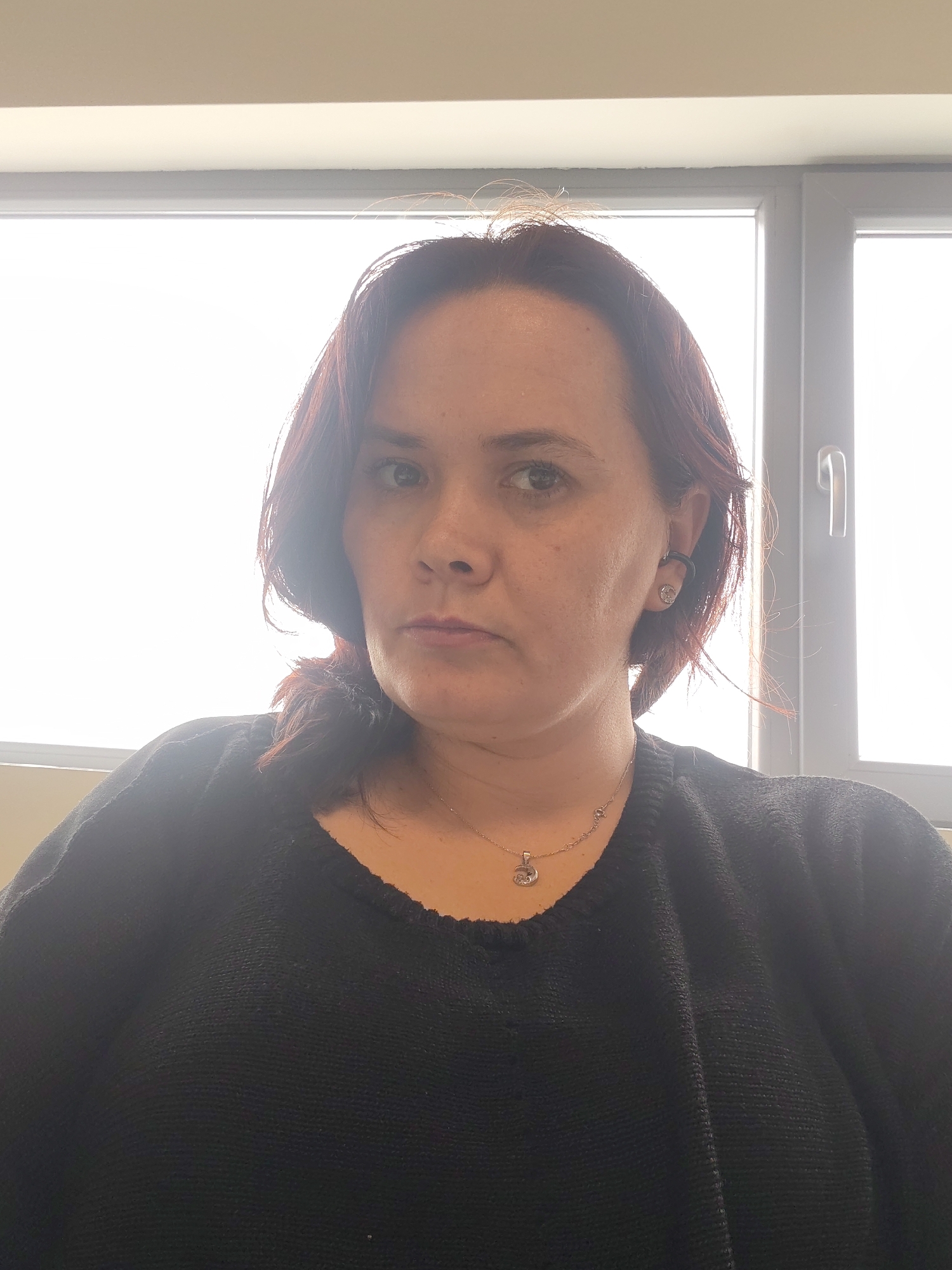 A woman with reddish-brown hair is facing the camera. She is wearing a black sweater and a necklace with a pendant. A window with a white frame is behind her. The location is not evident from the picture, therefore the location is undetermined.