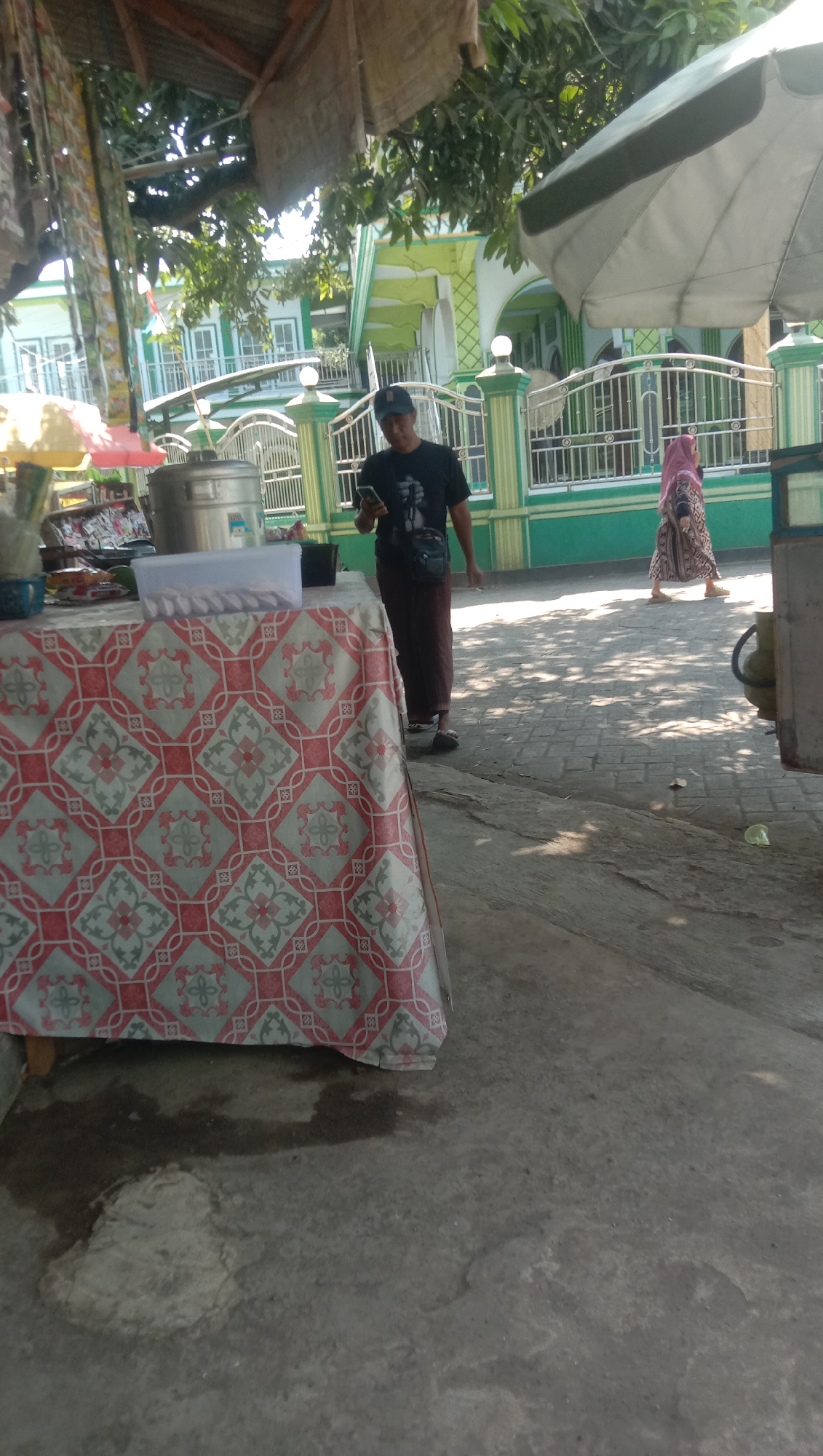The image depicts an outdoor scene in the city of Ngempit, Indonesia, likely near a place of worship. In the foreground, a table is covered with a red and white patterned tablecloth. On the table, there is a large metal container, a white plastic container with small items inside, and other assorted objects which are partially obscured. To the right of the table, a man wearing a dark t-shirt, brown pants, and a blue baseball cap is looking at his phone. He has a dark bag slung across his chest. Further in the background, a woman in a pink hijab and patterned dress is walking away from the camera. Behind them, a vibrant green and white building, possibly a mosque, is visible with arched windows and a fence in front of it. Trees with lush green leaves are present, casting shadows on the paved ground. The overall atmosphere suggests a sunny day.