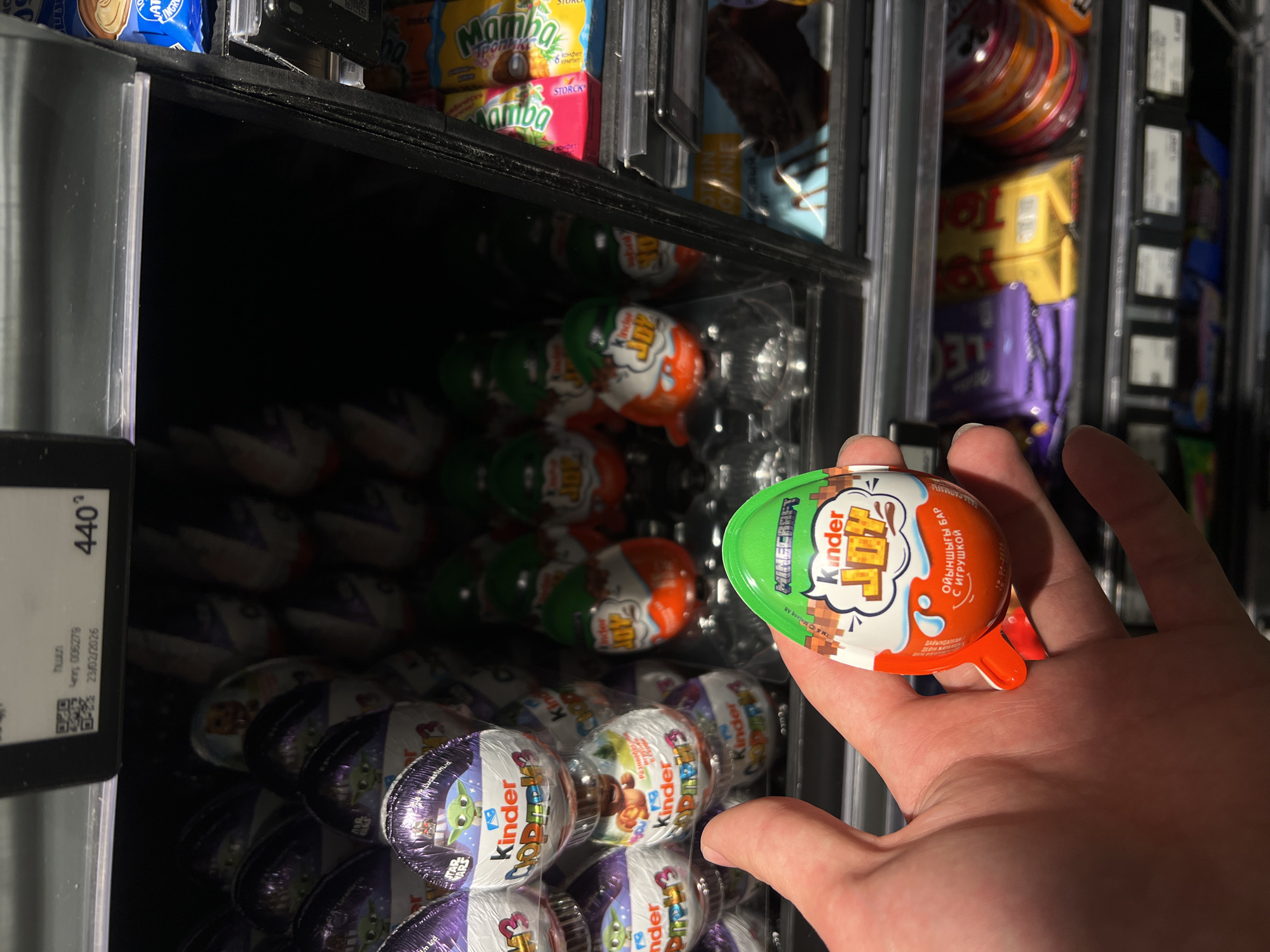 The image shows a person holding a Kinder Joy egg with Minecraft branding in a refrigerated display case, presumably in a store. The display case contains various Kinder Joy eggs with different themes, including Star Wars and other characters. Above the Kinder Joy eggs are Mamba candies and other products. A price tag on the left side of the display case shows "440" and a date of "23/02/2026". While the specific store and location in Yerevan, Armenia, cannot be determined from the image, it is likely a supermarket or convenience store selling these products.