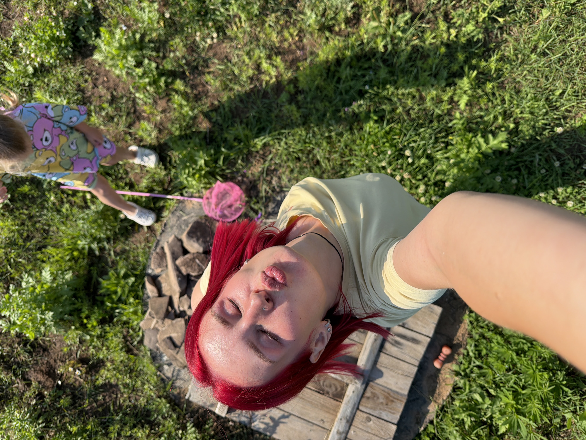 The image shows a top-down view of two people outdoors. In the foreground, a person with bright red hair is taking a selfie, puckering their lips and closing their eyes. They are wearing a light yellow t-shirt and a black choker necklace. A piercing is visible on their nose. In the background, a young child wearing a colorful t-shirt with a bear print is holding a pink butterfly net. The child is wearing white shoes. The scene is set in a grassy area with patches of dirt and some rocks. A wooden structure, possibly a bench or table, is visible on the right side of the frame. The sun is casting shadows, suggesting it is daytime, likely a sunny day. The overall atmosphere appears to be casual and outdoorsy, possibly in a backyard or park setting in Chaikovsky, Russia.