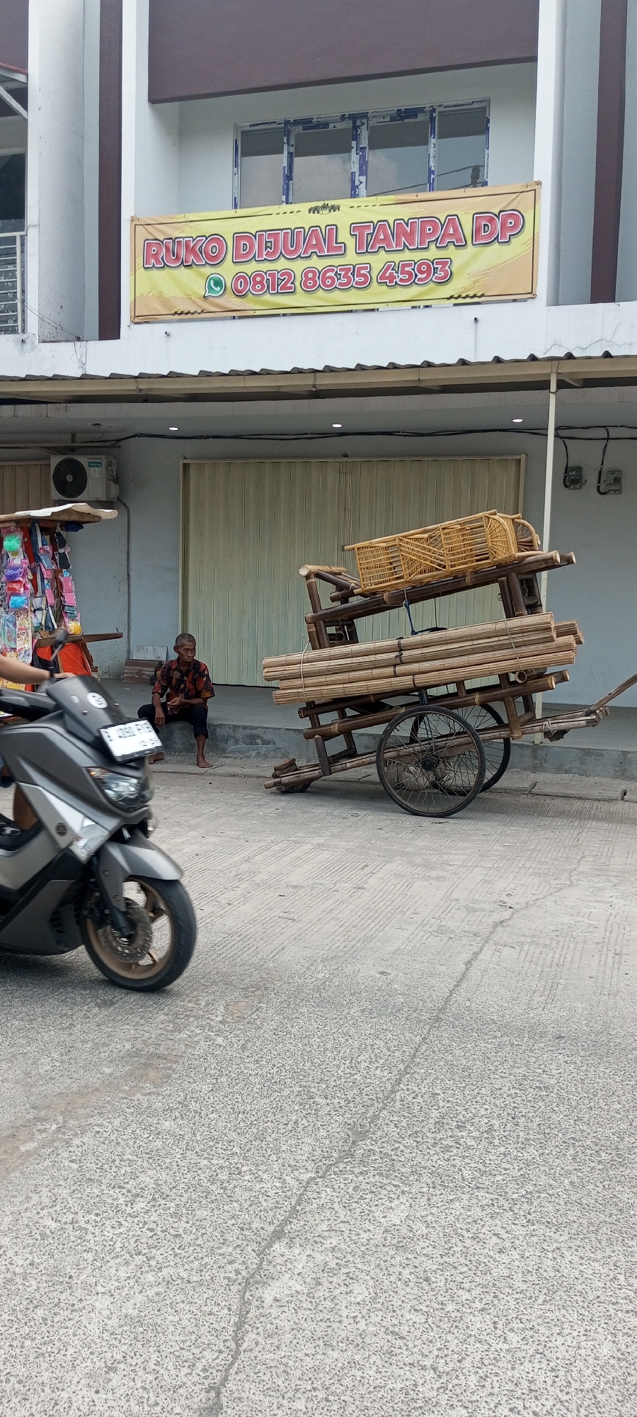 In Setu, Indonesia, a street scene depicts a man seated on a concrete step in front of a building. He is wearing a dark shirt with a red pattern and dark pants. To his right, a wooden cart is laden with bamboo poles and a woven basket. The cart has two large spoked wheels. In the foreground, a grey scooter is parked, facing the viewer. The building behind the man has a large yellow banner that reads "Ruko Dijual Tanpa DP 0812 8635 4593". The building has a metallic roll-up door and an air conditioning unit mounted on the exterior wall. A small stall with colorful items is visible to the left of the man. The street is paved with textured concrete.