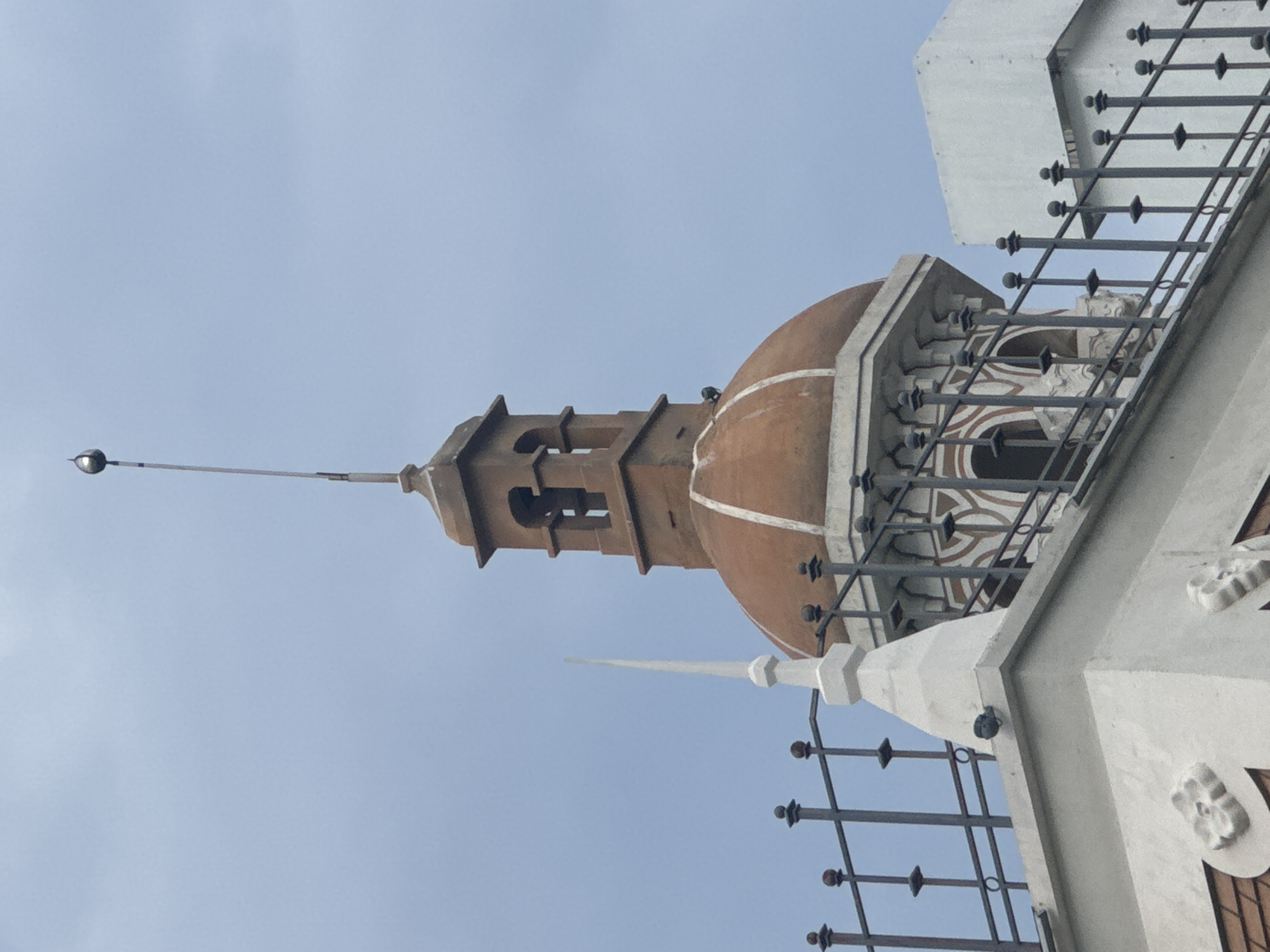 This is a close-up, low-angle shot of the spire and dome of a building against a cloudy sky. The spire is metallic and thin, topped with a bulbous object. Below the spire is a square, open bell tower structure. Underneath that is a rounded, reddish-brown dome with white trim. The dome sits atop an octagonal base with decorative white elements and arched openings. In the foreground, a dark metal fence with finials runs across the bottom of the frame. To the left of the dome, a white, pointed finial rises from a white architectural element. Part of a white rectangular building is visible to the right. The image does not contain enough information to determine the location in Nha Trang, Vietnam.