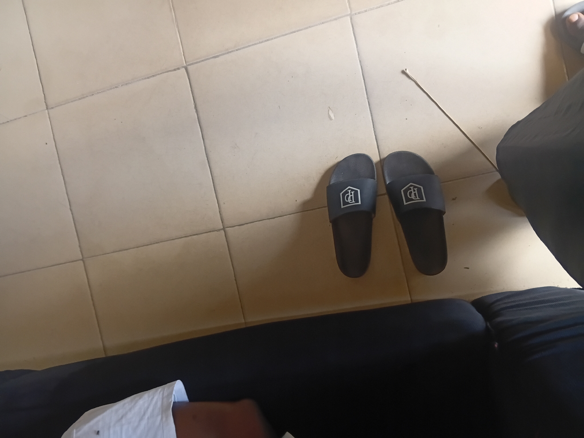 An overhead shot shows a pair of dark gray slip-on sandals with a white emblem on a beige tiled floor. The sandals are positioned horizontally, with the top sandal slightly above and to the left of the bottom sandal. To the left of the sandals, a black sofa is visible, and a person's legs, clad in dark pants, are partially in view at the bottom right of the frame. A thin, white object, possibly a straw or a thin stick, lies diagonally across the floor between the sandals and the person's legs. The scene appears to be indoors, likely in a residential setting in Jalingo, Nigeria, as indicated by the beige tile flooring commonly found in homes in the region. The lighting is soft, suggesting it could be daytime.
