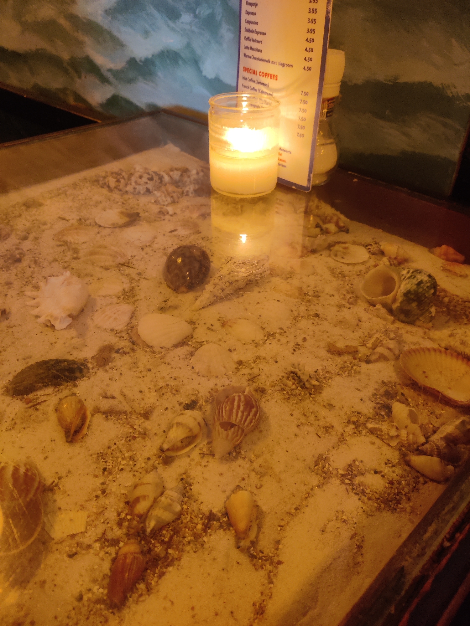 The image depicts a close-up view of a glass-topped table filled with sand and seashells. A lit white candle in a glass jar is positioned on the left side of the frame. To the right of the candle, a menu with Dutch text and prices is propped up, accompanied by a salt and pepper shaker. The table surface is adorned with various seashells, some whole and some broken, interspersed with grains of sand. The background features a painting or mural with abstract wavy patterns in shades of green and blue. The scene appears to be set indoors, possibly in a cafe or restaurant in The Hague, Netherlands, given the presence of the menu with Dutch language. There are no people visible in the image.
