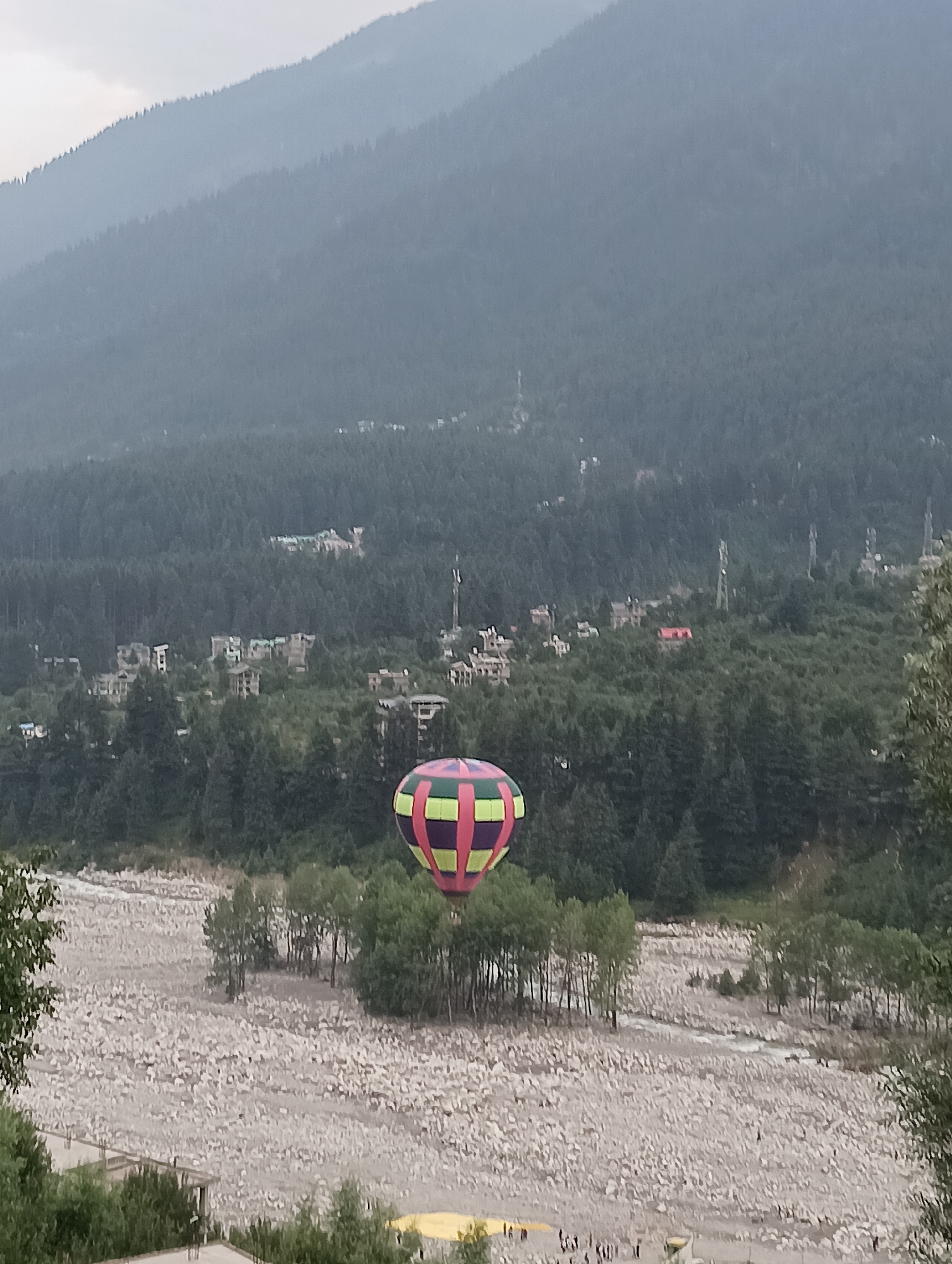 A hot air balloon, with a colorful pattern of pink, green, and yellow panels, is visible in the lower right of the image. It is ascending over a rocky riverbed, which is dry and filled with a dense collection of light-colored stones. Scattered trees grow in patches throughout the rocky terrain. In the background, a lush, green forest covers the slopes of a large mountain. Several buildings, appearing to be houses or small hotels, are nestled within the treeline. The sky is overcast and hazy, suggesting either early morning or late afternoon. The overall impression is a serene, natural landscape with a touch of adventure offered by the hot air balloon. There is no visible text in the image.
