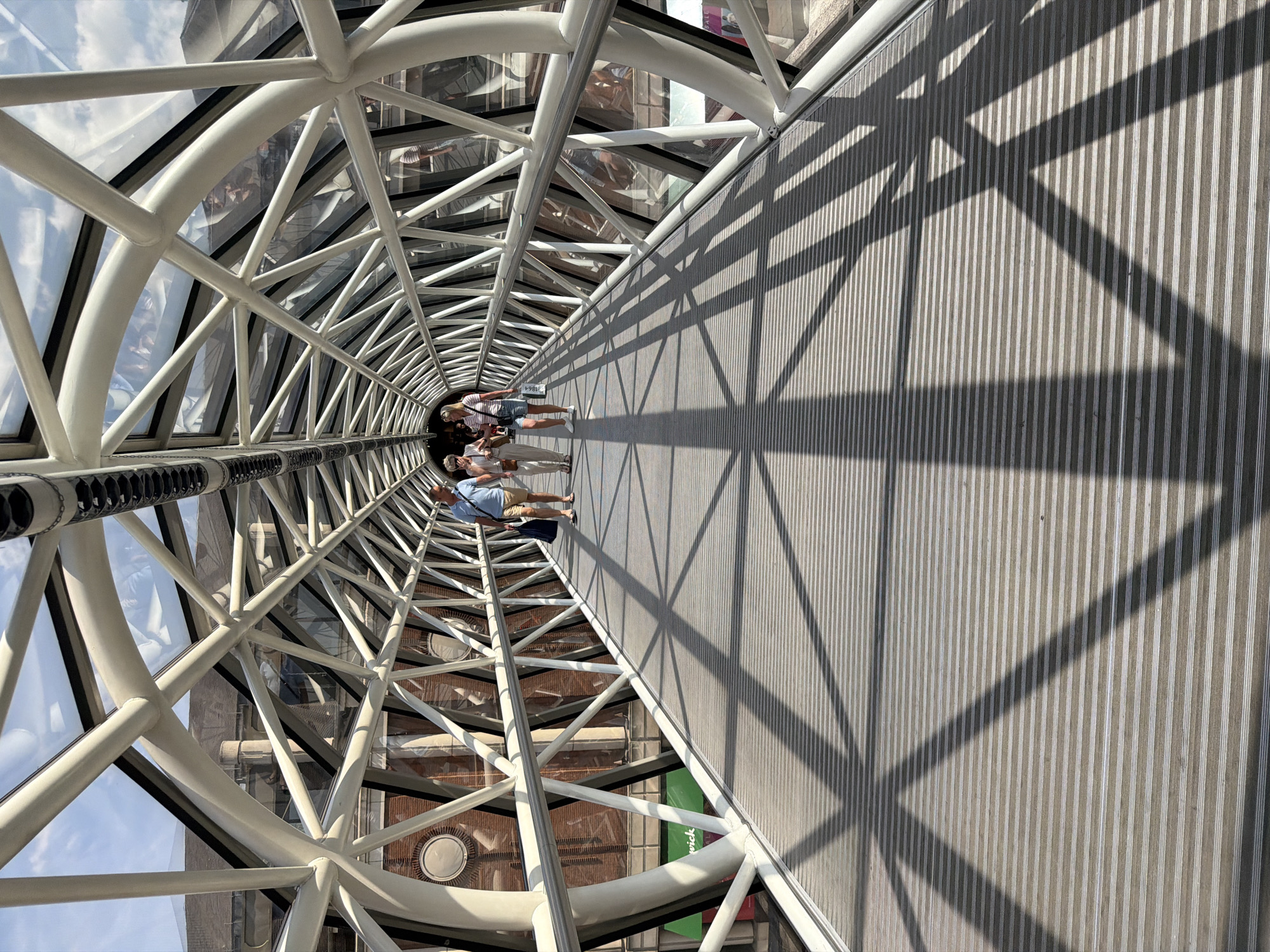 This image captures a pedestrian walkway inside a modern, glass-enclosed bridge with a white tubular metal framework. The bridge is covered by a transparent roof and sides, allowing natural light to illuminate the interior. The floor of the walkway is made of grey, ribbed material, and the shadow of the bridge's structure is cast across it in a geometric pattern. Three people are walking along the bridge away from the camera. The person in the foreground on the left is a man wearing a blue shirt and light-colored shorts, carrying a dark bag. To his right are two women, one in a white top and patterned skirt, and the other in a white striped top and denim shorts. They are all walking casually. Through the glass sides of the bridge, a red brick building with circular window details and a black roof is visible, suggesting an urban environment. A green sign with white text is partially visible on the exterior wall of the building. The overall scene depicts a bright, sunny day in a city. This structure is likely the Millennium Bridge in London, UK, known for its modern design and pedestrian use.