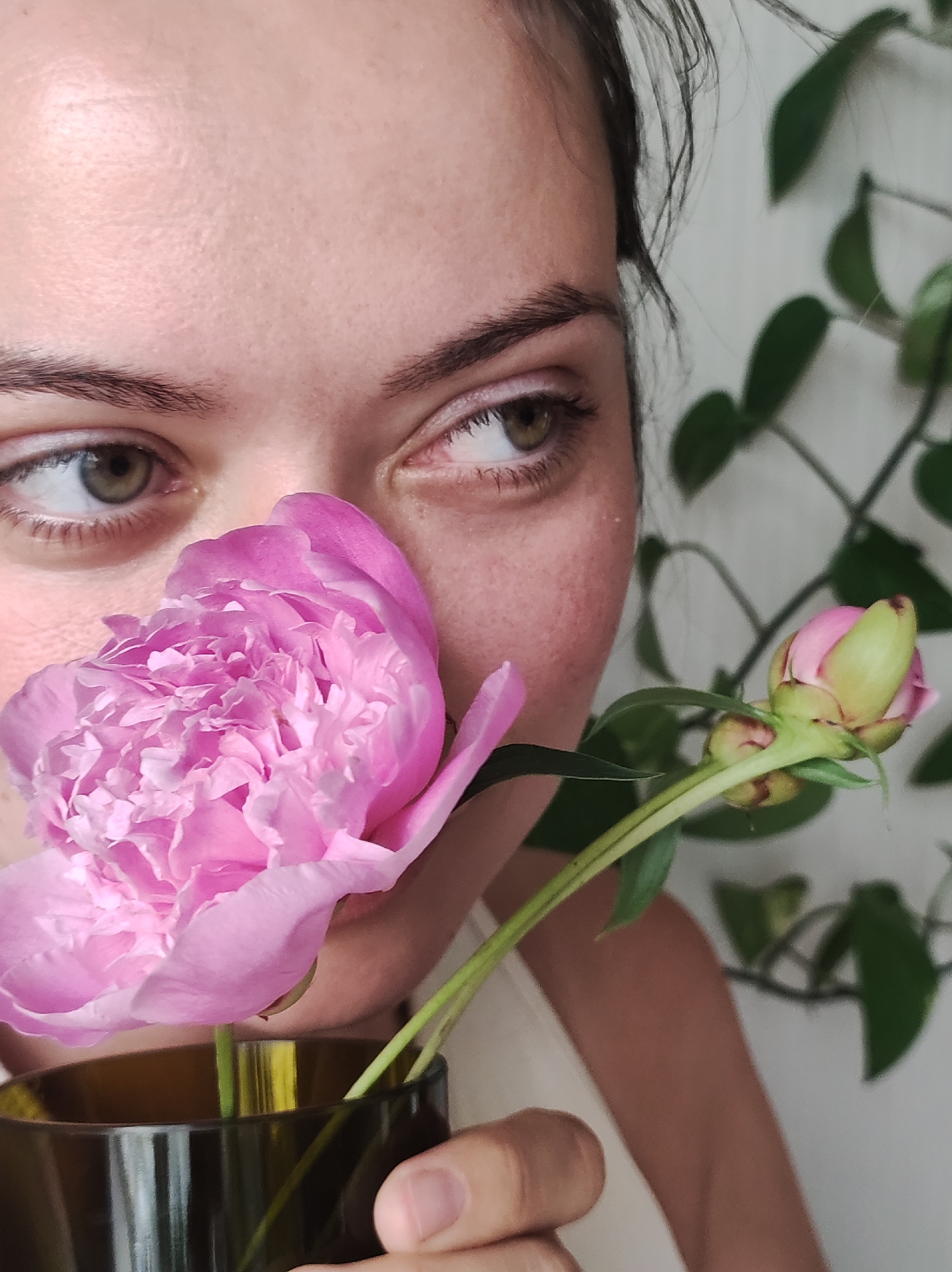 A close-up shot focuses on the lower face and right eye of a young woman, partially obscured by a large, fluffy pink peony flower. Her green eyes, with subtle pink eyeliner, gaze towards the right. A smaller peony bud, also pink and green, is visible to the right of the main flower. The woman holds a dark brown vase containing the flowers. Behind her, out of focus, is a green plant with large leaves and a light-colored wall. The image does not contain any elements that would indicate a specific location within the city of Chaikovsky, Russia.