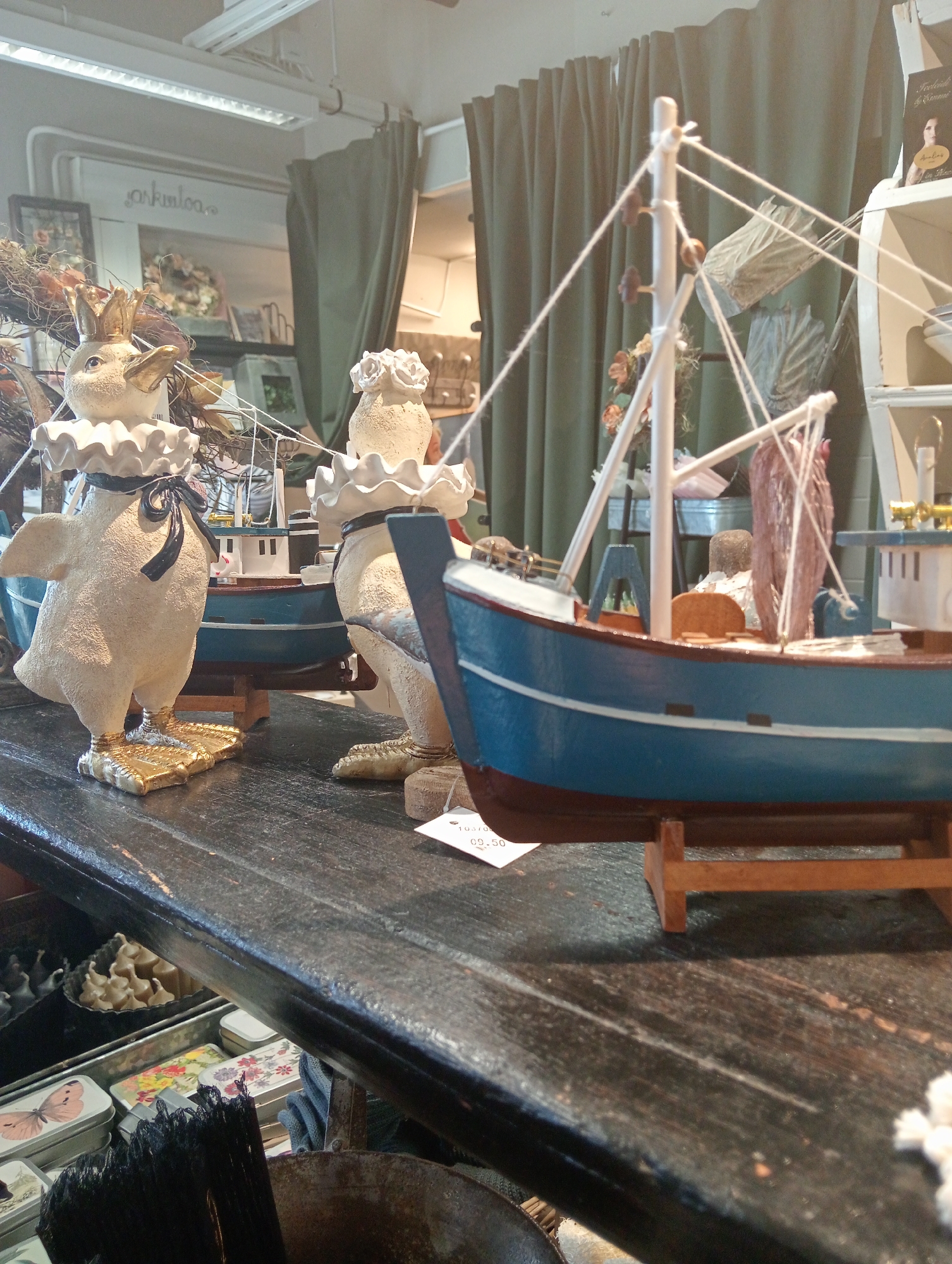 The image shows a shop interior in Kuopio, Finland, featuring a display of decorative items. In the foreground, two ceramic duck figures with ruffled collars and one wearing a crown are positioned on a dark wooden surface. Behind the ducks, a model of a blue and white fishing boat is prominently displayed on a wooden stand. The background reveals a green curtain and shelves stocked with various items, including framed pictures and decorative elements. Several metal tins with floral and butterfly patterns are visible on the lower shelf of the wooden surface. The overall scene suggests a retail environment selling home decor or gift items.