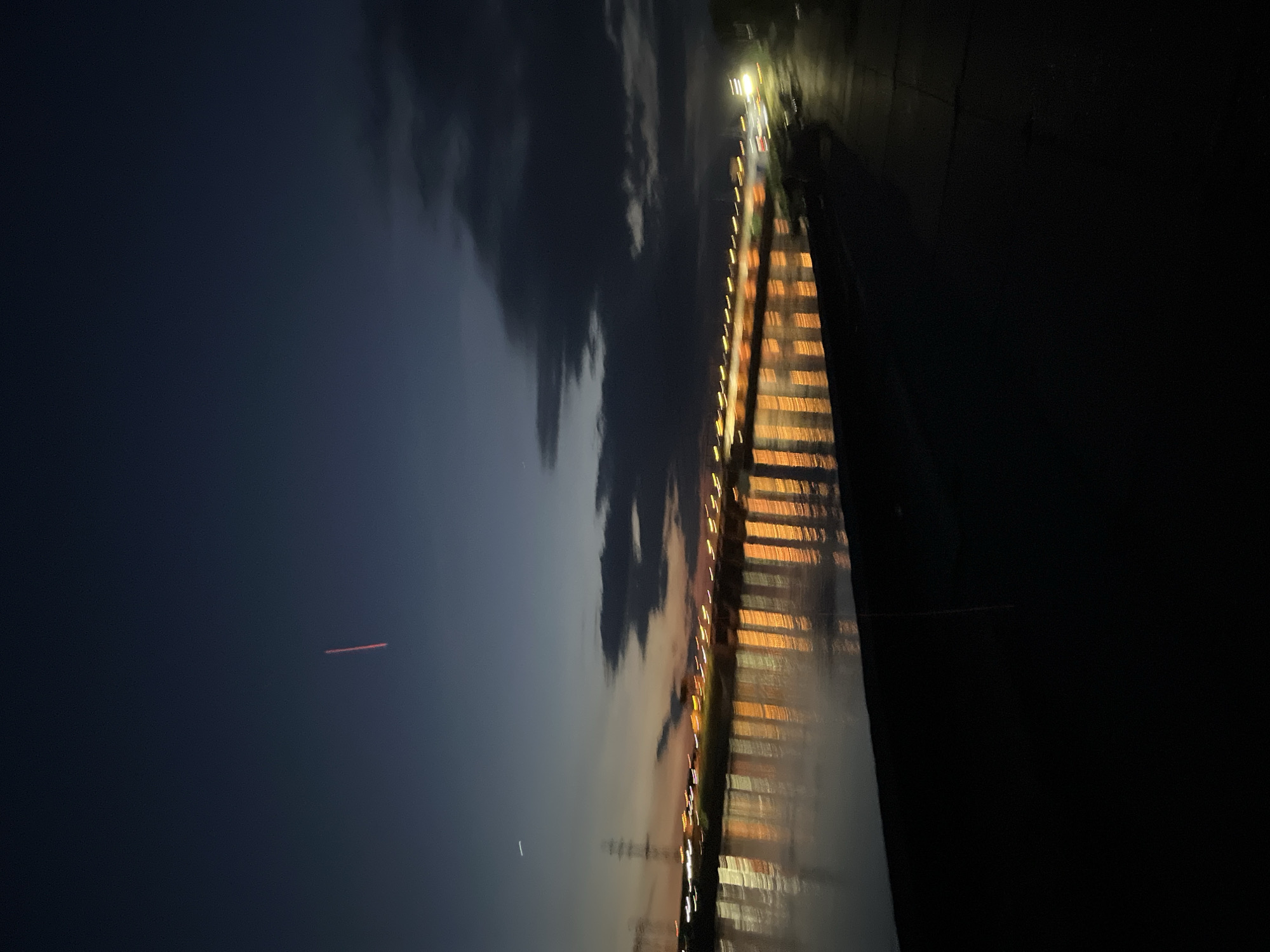 The image captures a serene nighttime waterscape in Chaikovsky, Russia, likely along the Kama River. The scene is dominated by a dark blue sky with scattered clouds, indicating late dusk or night. A long structure, possibly a pier or a section of a bridge, extends horizontally across the middle ground. Its side facing the viewer is lined with numerous rectangular lights, casting warm orange and yellow reflections that shimmer vertically on the dark water below.
At the far end of this illuminated structure, a bright streetlamp-like light illuminates a section of what appears to be a paved area or roadway, with other fainter lights visible further in the distance, suggesting a city or built-up area along the water's edge. In the dark expanse of the sky, a faint, reddish streak of light is visible, possibly an airplane contrail or a meteor, along with another much fainter white streak. The silhouettes of what appear to be power lines or transmission towers are faintly discernible on the horizon. The overall atmosphere is peaceful and calm, with no people visible. There is no visible text in the image.