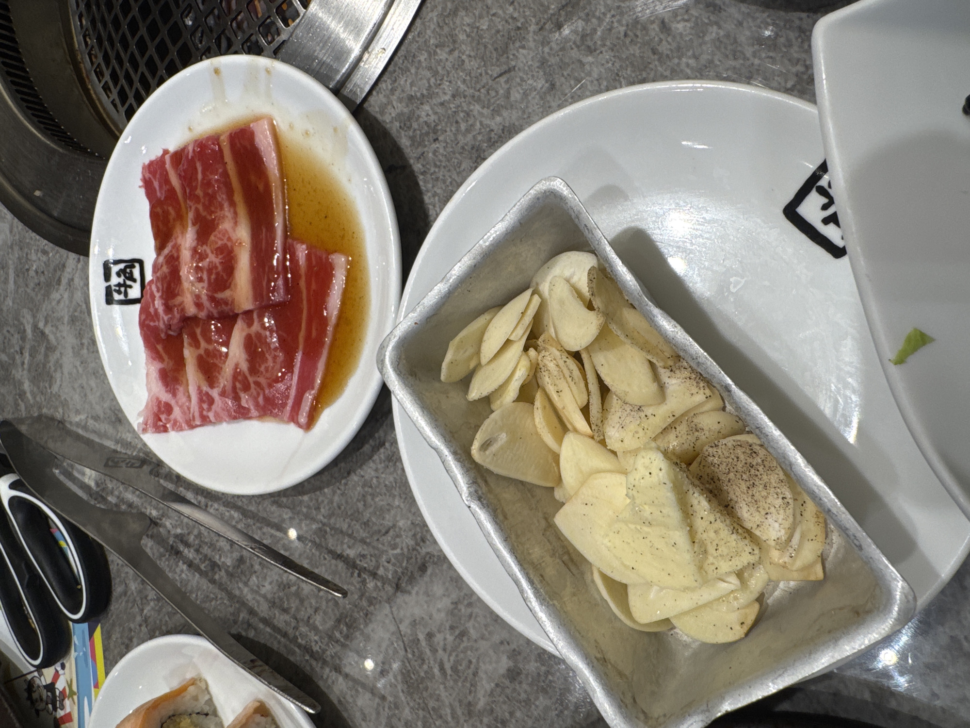 The media file captures an overhead view of a dining table setting, likely in a restaurant in Kuta Selatan, Indonesia, showcasing ingredients prepared for a meal.
In the upper left, a white circular plate holds multiple slices of raw, marbled red meat, possibly beef or pork belly, soaking in a light brown marinade or sauce. This plate features a black square logo with a white, stylized character inside, resembling a Kanji or Hanzi character. Adjacent to it, a rectangular metal container is filled with numerous thin slices of garlic, visibly seasoned with coarse black pepper, suggesting they are intended for grilling or cooking. This metal dish rests on a larger, plain white plate, which also displays a less distinct black, stylized character logo near its edge.
A pair of gleaming metal tongs lies on the grey, speckled table surface, positioned below the meat plate and to the left of the garlic, indicating their use for handling food during cooking. Partially visible in the lower left corner, a white bowl contains what appears to be segments of sushi rolls, hinting at a diverse menu. The edge of a circular metal grill or hot plate is discernible in the upper left background, suggesting a Korean BBQ or Japanese Yakiniku dining experience.
The scene depicts the fresh ingredients laid out before the cooking process begins, under ambient indoor lighting. The visible text consists of the stylized black and white logos on the plates, likely restaurant branding.