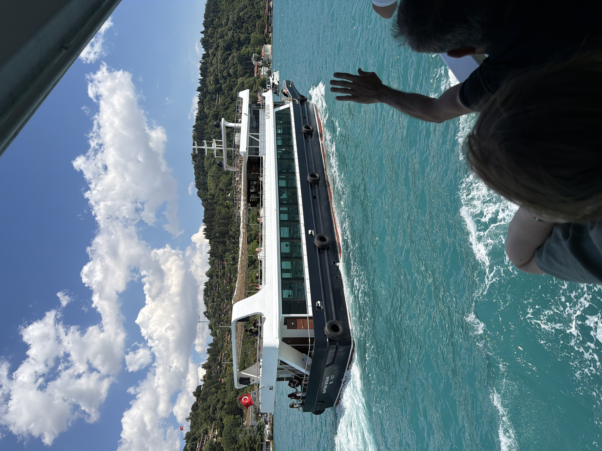 A large, dark blue passenger ferry named "KAPTAN BORAN BL IST" sails through bright turquoise water under a partly cloudy blue sky. The ferry has a white upper deck with patrons visible. A Turkish flag flies from the front of the upper deck. The ferry is leaving a wake behind it. On the right side of the image, the arm and hand of a person are visible, waving towards the ferry. In the background, a lush green, tree-covered hillside with houses is visible. The scene is likely the Bosphorus Strait, near Üsküdar, Turkey.