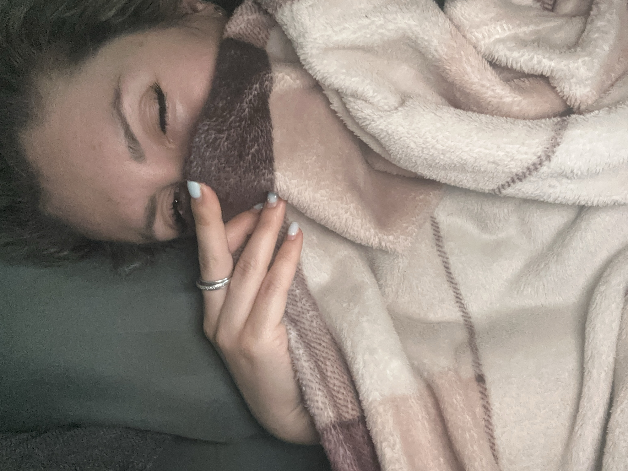 A close-up shot captures a woman, partially obscured by a soft, patterned blanket, as she lies on a dark-colored surface, likely a couch. Her right eye is visible, closed in a wink, while her left eye glances forward. Her hand, adorned with a silver ring, rests on the blanket, with light blue nail polish on her fingernails. The blanket features a plaid pattern with shades of brown, pink, and white. The background is a plain, dark green, suggesting an indoor setting. The image is taken from a high angle, looking down at the woman. The location of this scene within Novoaltaysk, Russia, cannot be determined from the visual information provided.