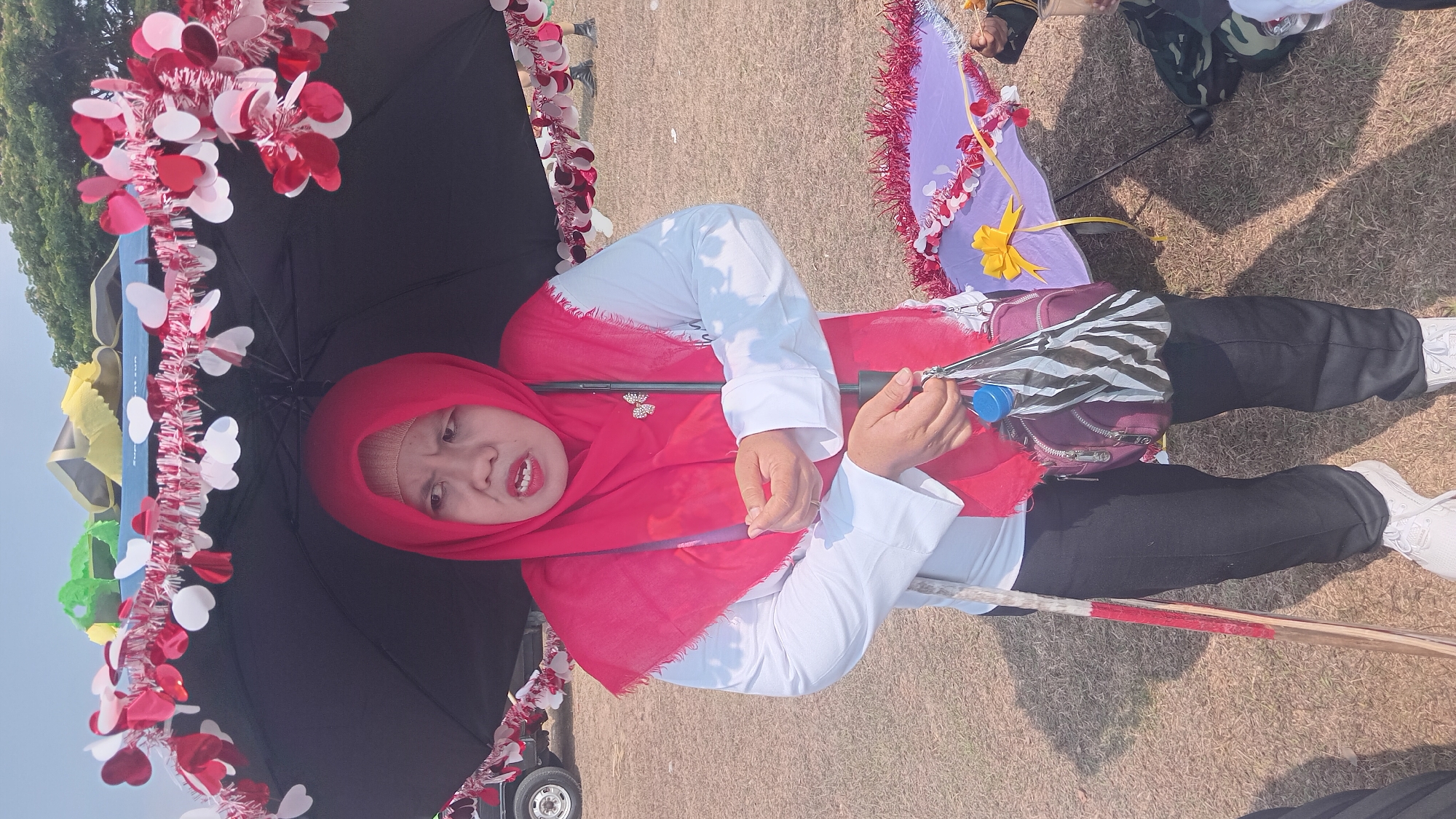 A woman wearing a red hijab and white shirt stands under a decorated black umbrella. She is holding a small purple bag and a folded umbrella. To her left, a wooden pole with red and white stripes is visible. In the background, tents and trees are present. The scene appears to be an outdoor event. The image is a medium shot, focusing on the woman and her immediate surroundings. The overall lighting suggests it is daytime.