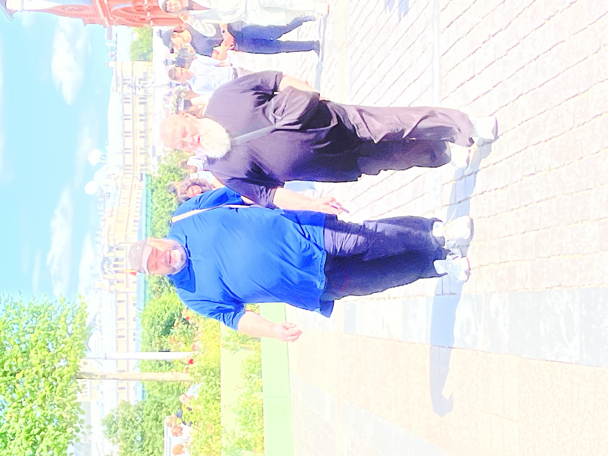 Two men walk on a paved pathway in Moscow, Russia. The man on the left wears a blue polo shirt and dark pants, with a baseball cap on his head. The man on the right wears a dark t-shirt, dark pants, and carries a dark messenger bag over his shoulder; he has a white beard and is wearing glasses. Behind them, other pedestrians are visible, as well as trees and buildings in the background, indicating an outdoor public space, possibly a park or a street. The sky is bright blue with some clouds.