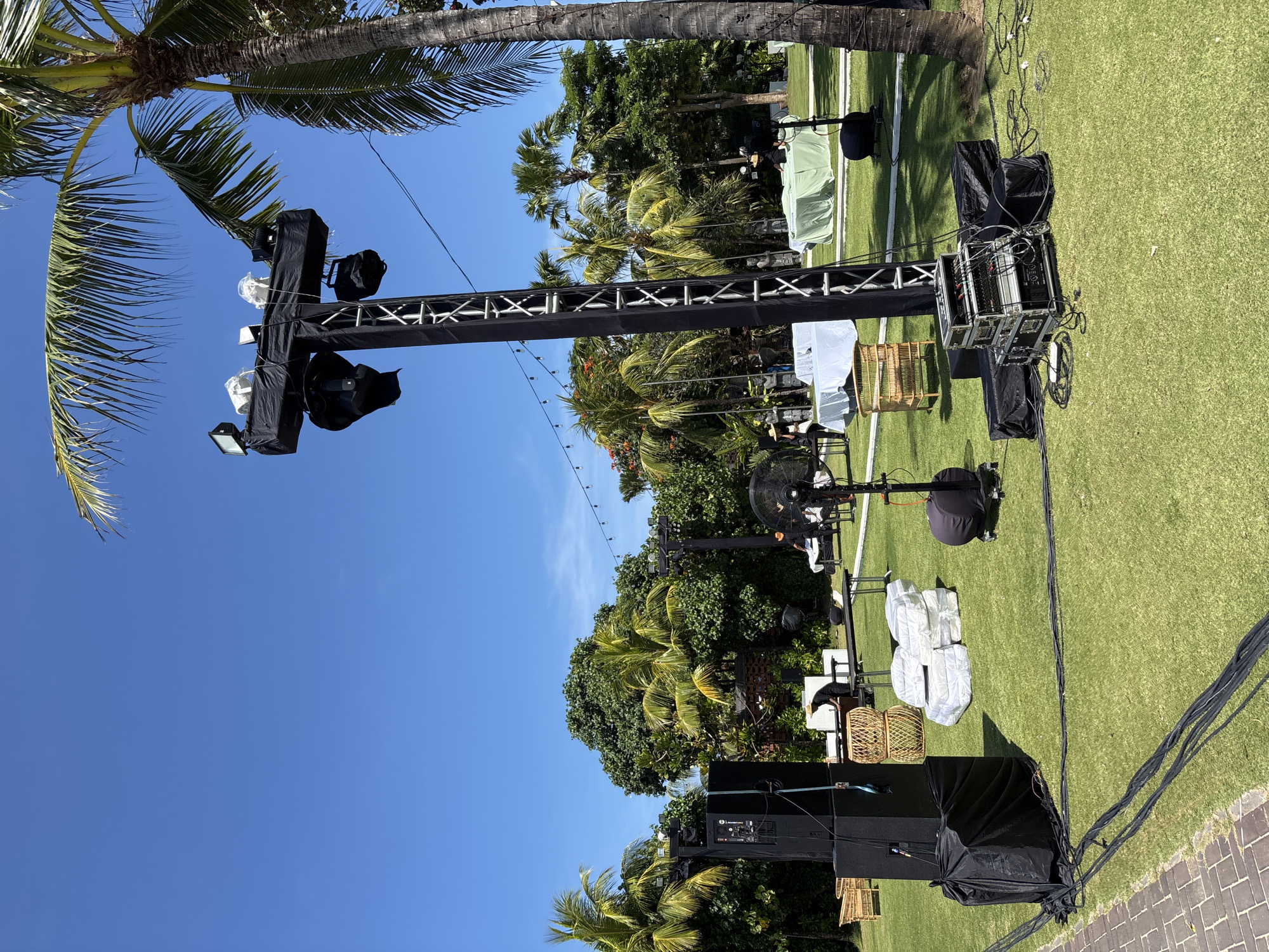 The image shows a grassy outdoor area under a clear blue sky, possibly in Kuta Selatan, Indonesia, given the presence of palm trees and tropical foliage. A tall, black metal lighting truss stands prominently, with several covered lights and loudspeakers attached. Cables are visible, running along the ground. Miscellaneous objects, including covered chairs and wicker baskets, are scattered on the grass. Some people are present.