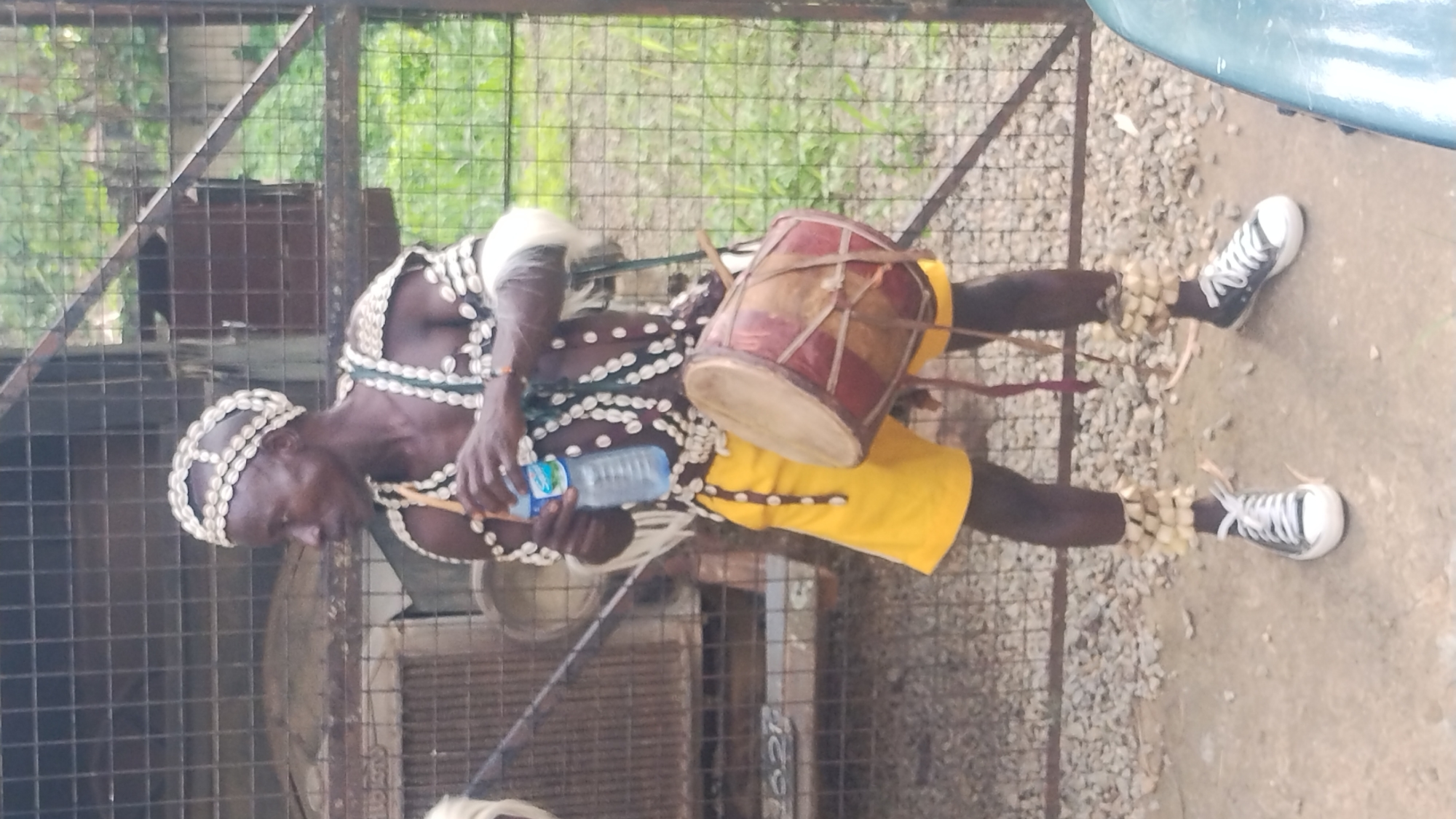 The image shows a person adorned in a traditional outfit, possibly a performer or participant in a cultural event, in Jos, Nigeria. The subject is wearing a headdress and attire decorated with white shells, a beaded necklace, and a yellow garment. They are holding a bottle of water and a small drum, suggesting they might be about to perform or have just finished.
The scene appears to be outdoors, with a metal fence in the background and gravel on the ground. The lighting suggests it is daytime, and the background foliage indicates a possibly warm climate. The subject's expression is neutral, and their posture is relaxed, with one hand holding the water bottle and the other likely resting on the drum.
Notable details include the traditional shell adornments, the drum, and the modern sneakers worn by the subject, creating an interesting juxtaposition of traditional and contemporary elements. The presence of a metal fence and what appears to be a concrete or dirt ground suggests an urban or semi-urban environment within Jos, Nigeria. There is no visible text in the image. The overall impression is of a cultural presentation or a moment of preparation for one.
