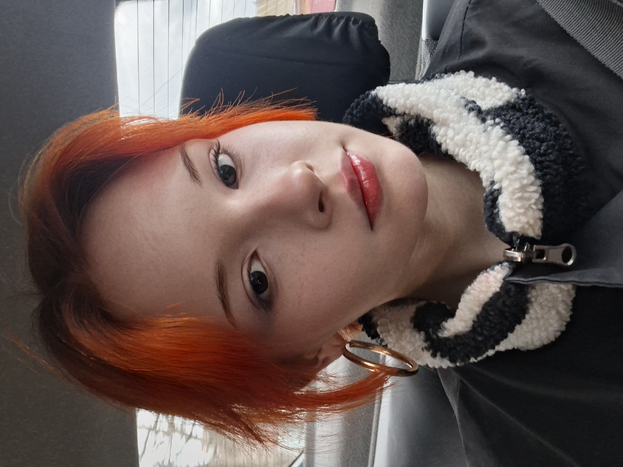 A selfie taken inside a vehicle showing a person with bright orange hair, a black and white jacket with a sherpa collar, and gold hoop earrings. The location is Chaikovsky, Russia, inferred from the vehicle's window reflection, which shows a blurred background resembling urban features with a high-rise building. The person is positioned at an angle, facing the camera. They are wearing red lipstick. The background includes a seat and a window with a reflection.