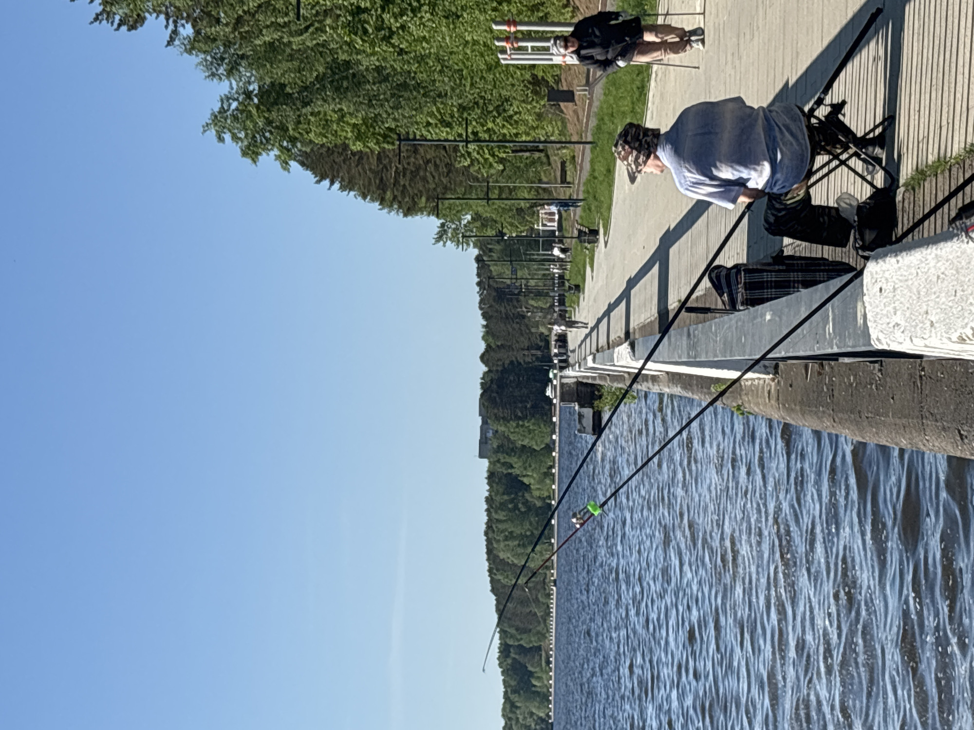 A man is fishing from a concrete embankment in Chaikovsky, Russia. He is wearing a light blue shirt and a camouflage hat, and he is sitting in a black folding chair. Fishing rods extend out over the water. Another person is visible in the background. The embankment has black metal railings. Trees line the opposite shore. The sky is clear and blue.