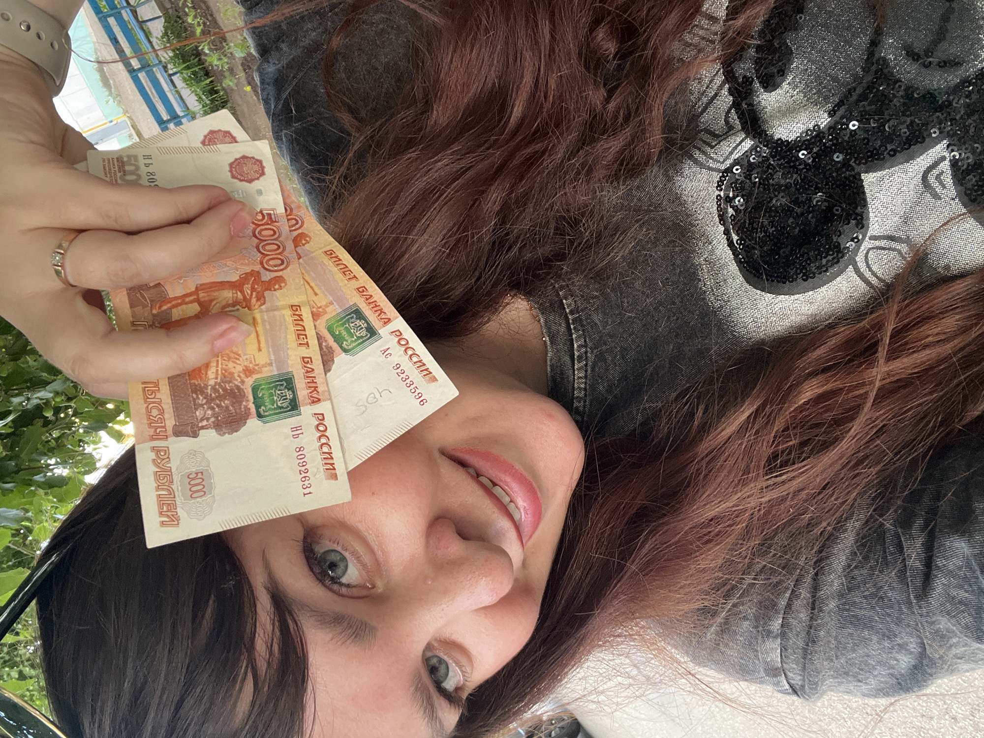 A female individual is depicted in a close-up, upward-angle selfie, reportedly taken in the city of Chaikovsky, Russia. The individual has long, wavy, reddish-brown hair and light-colored eyes. She is smiling, revealing her teeth. She wears a dark, textured garment, possibly denim, featuring a black sequined design over her right shoulder. Her right hand holds three 5000 Russian Ruble banknotes vertically near her face. Partially visible serial numbers on two notes include 'НВ 8092631' and 'AC 9233596'. A gold-toned ring with two small stones is present on her right ring finger. A light-colored smartwatch is partially visible on her left wrist. The background features green foliage, with a blue-railed structure and a white, translucent structure, possibly a greenhouse, discernible in the distance.