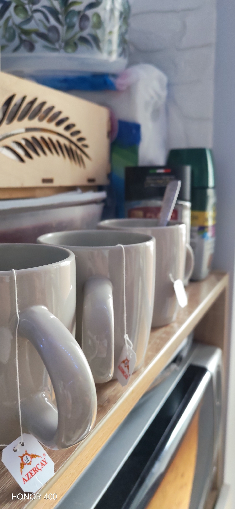 Three light-grey mugs sit on a wooden shelf above a microwave. Each mug has a tea bag dangling from its handle, with the tag on the leftmost mug displaying the brand name "AZERCAY" in orange lettering. A wooden box with a fern-like cutout pattern sits behind the mugs, along with various other containers and jars. The image includes the watermark "HONOR 400" at the bottom left.