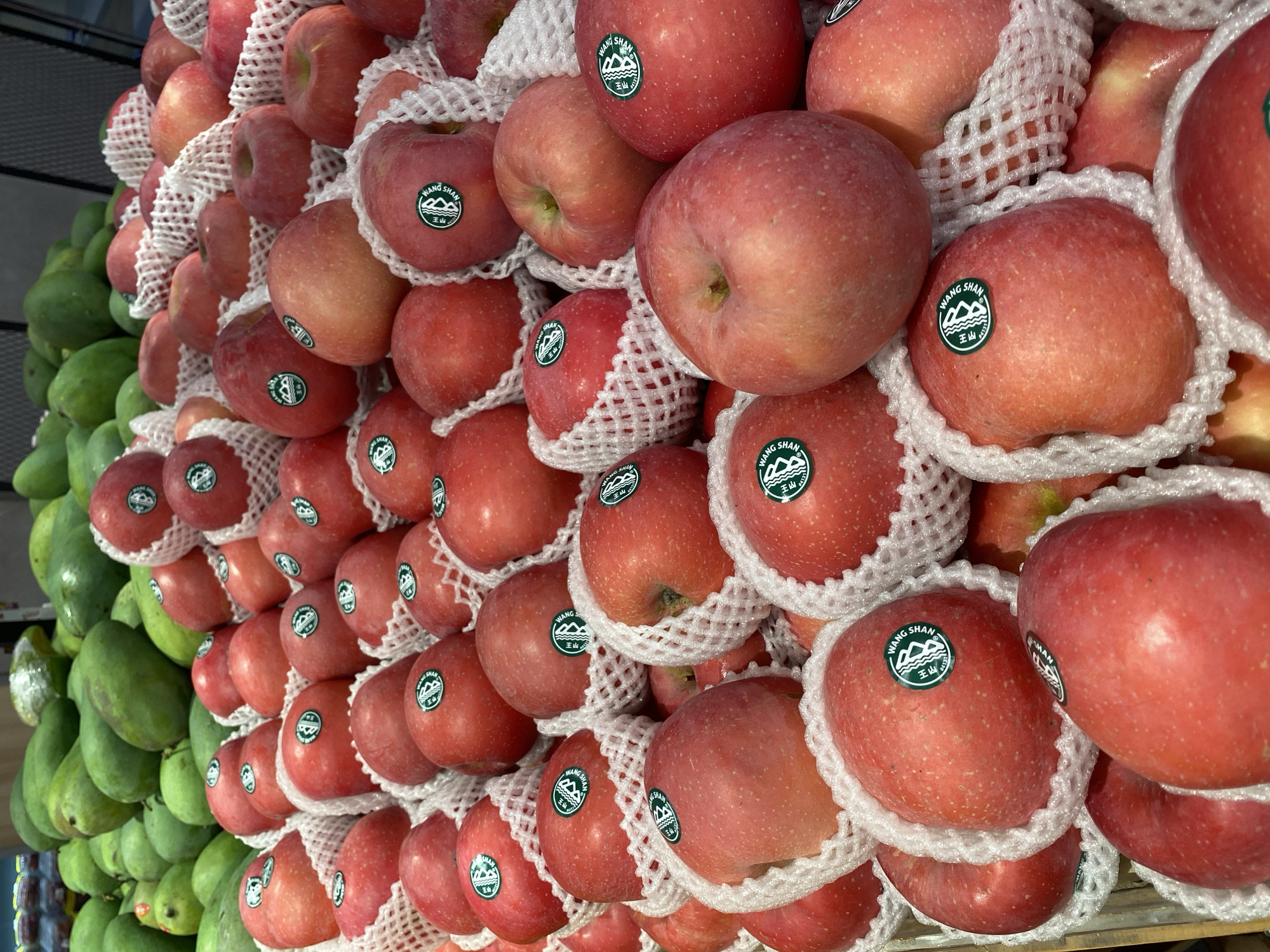 The image shows a close-up view of a pile of red apples. The apples are individually wrapped in white foam netting and arranged in plastic trays. Many of the apples have round green stickers with Chinese characters and the words "WANG SHAN" printed on them. In the background, out of focus, is a pile of green mangoes. The lighting suggests an indoor setting, likely a market or grocery store. The scene is devoid of people. The image captures a commercial display of produce, with a focus on the apples and their packaging. The specific location in East Jakarta, Indonesia, is not evident from the visual information alone.