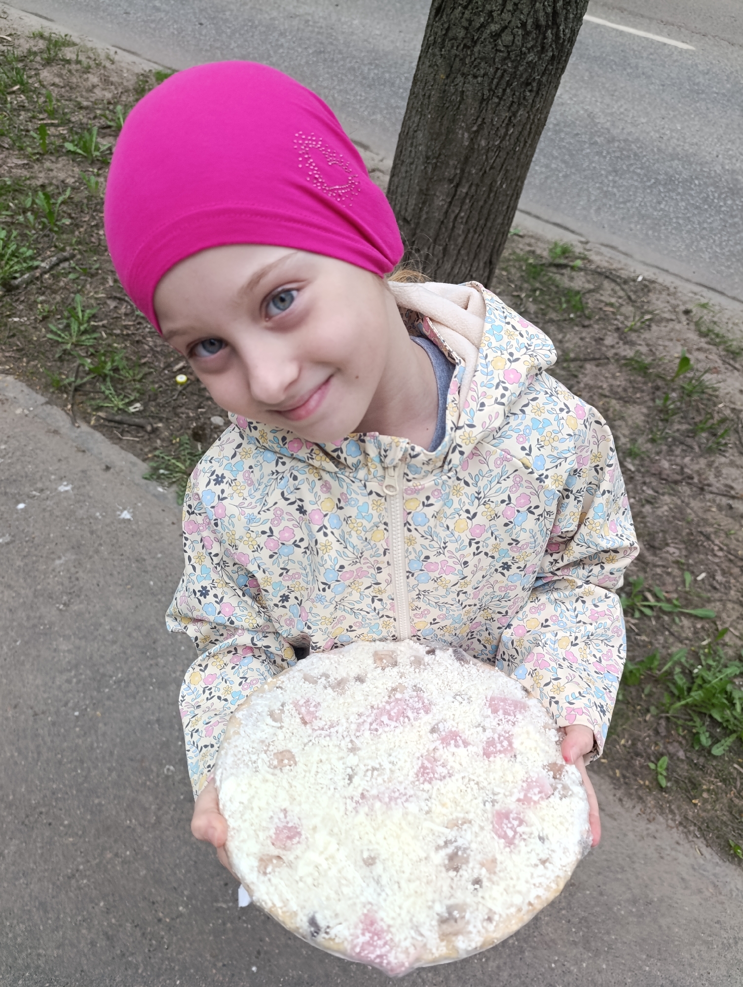 A young girl with blue eyes and a pink headscarf is holding a pizza with a white, cheese-covered topping. She is wearing a floral-patterned hooded jacket. The background includes a tree trunk and pavement. The location appears to be an outdoor area near a street in the city of Unknown, Unknown.